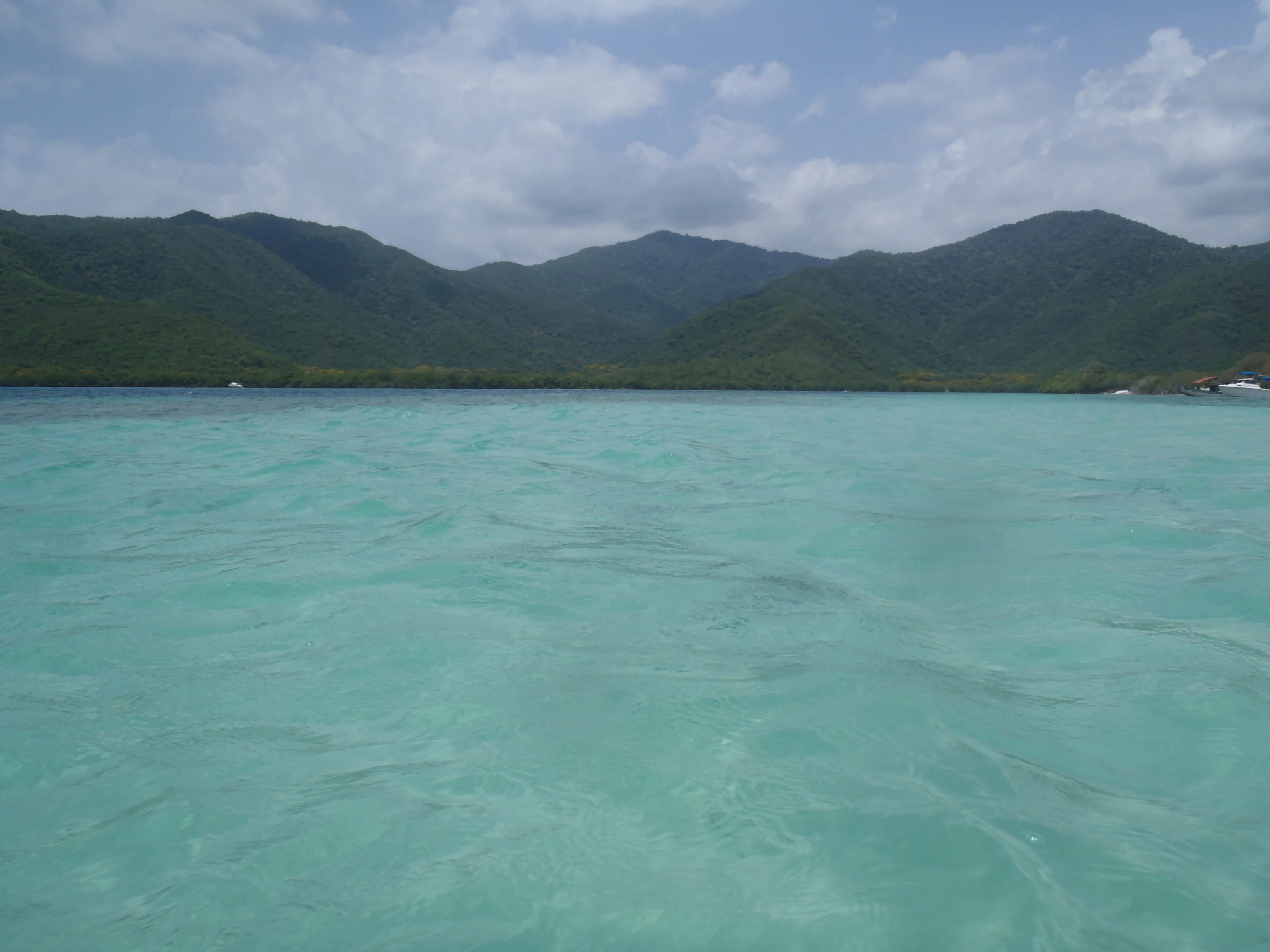 Turquoise water with gentle waves extending toward mountainous coastline under partly cloudy sky