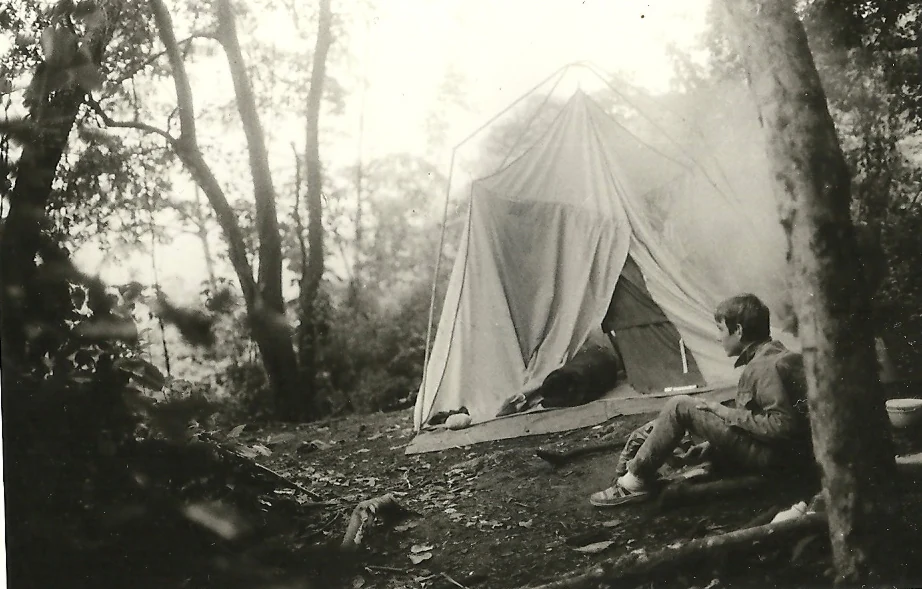 Black-and-white photograph of a person sitting next to a canvas tent in a forested area with trees and foliage surrounding the campsite