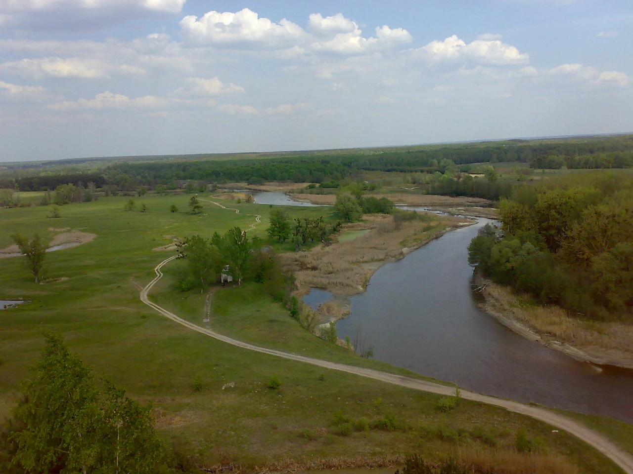 Wide landscape view of a river flowing through grassy fields with a winding path, trees, and open sky