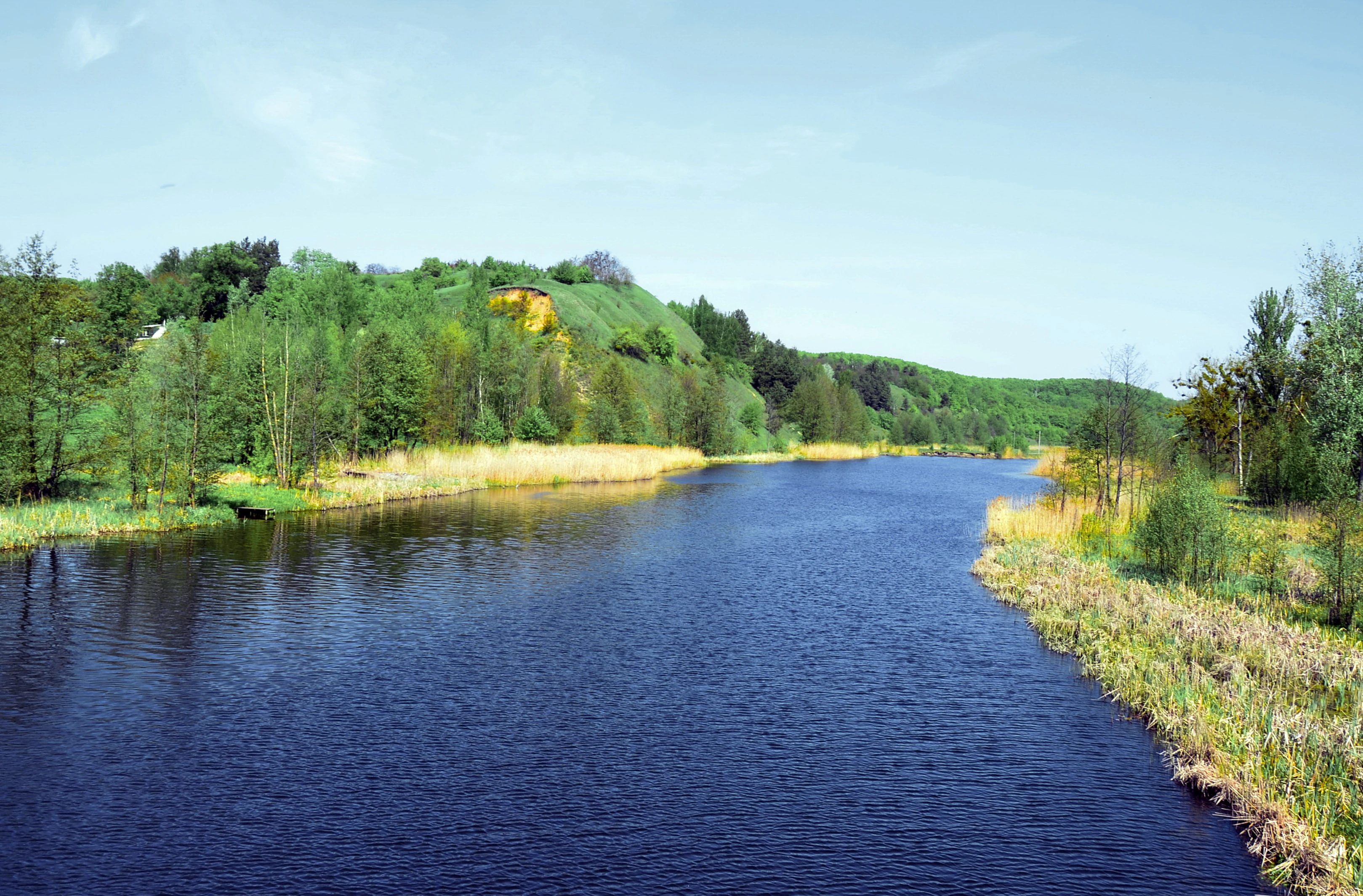 A wide river with dark blue water flows through a landscape with green vegetation and hills under a clear sky.