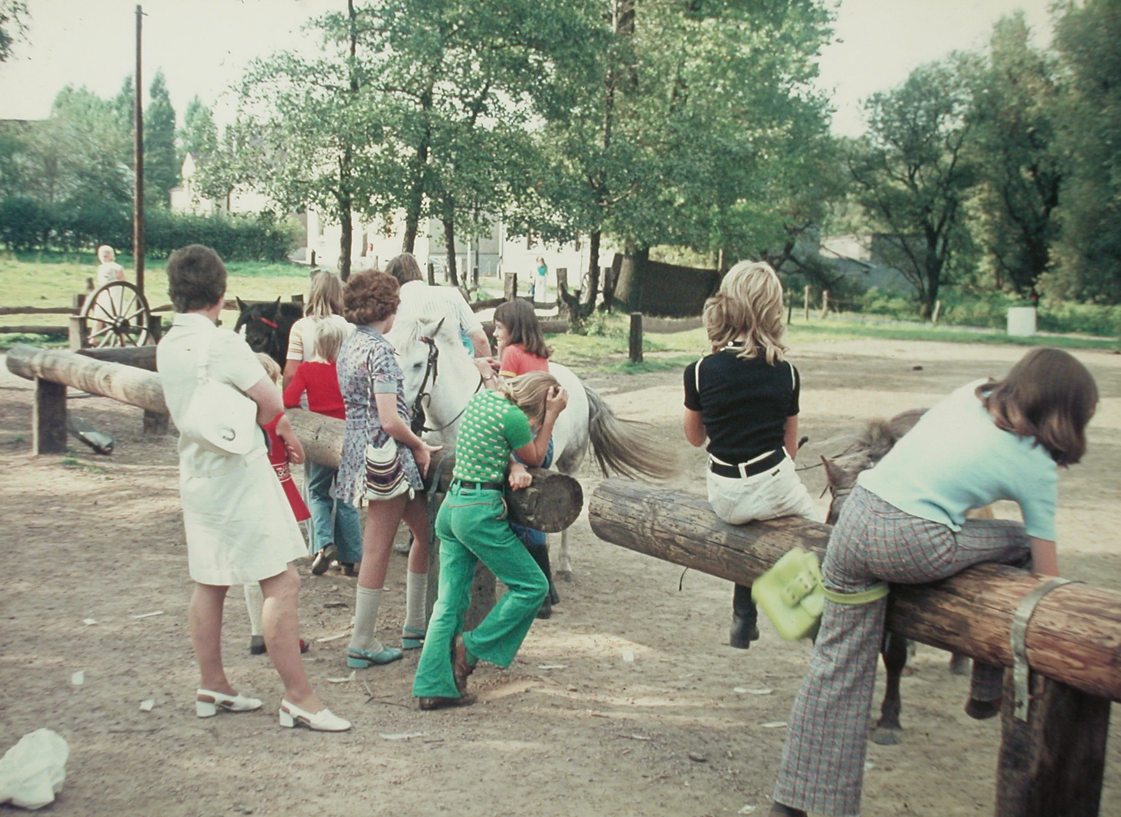 A group of people, mostly children and young adults, gathered at an outdoor equestrian area in a rural setting, with several sitting on a heavy log fence near ponies.