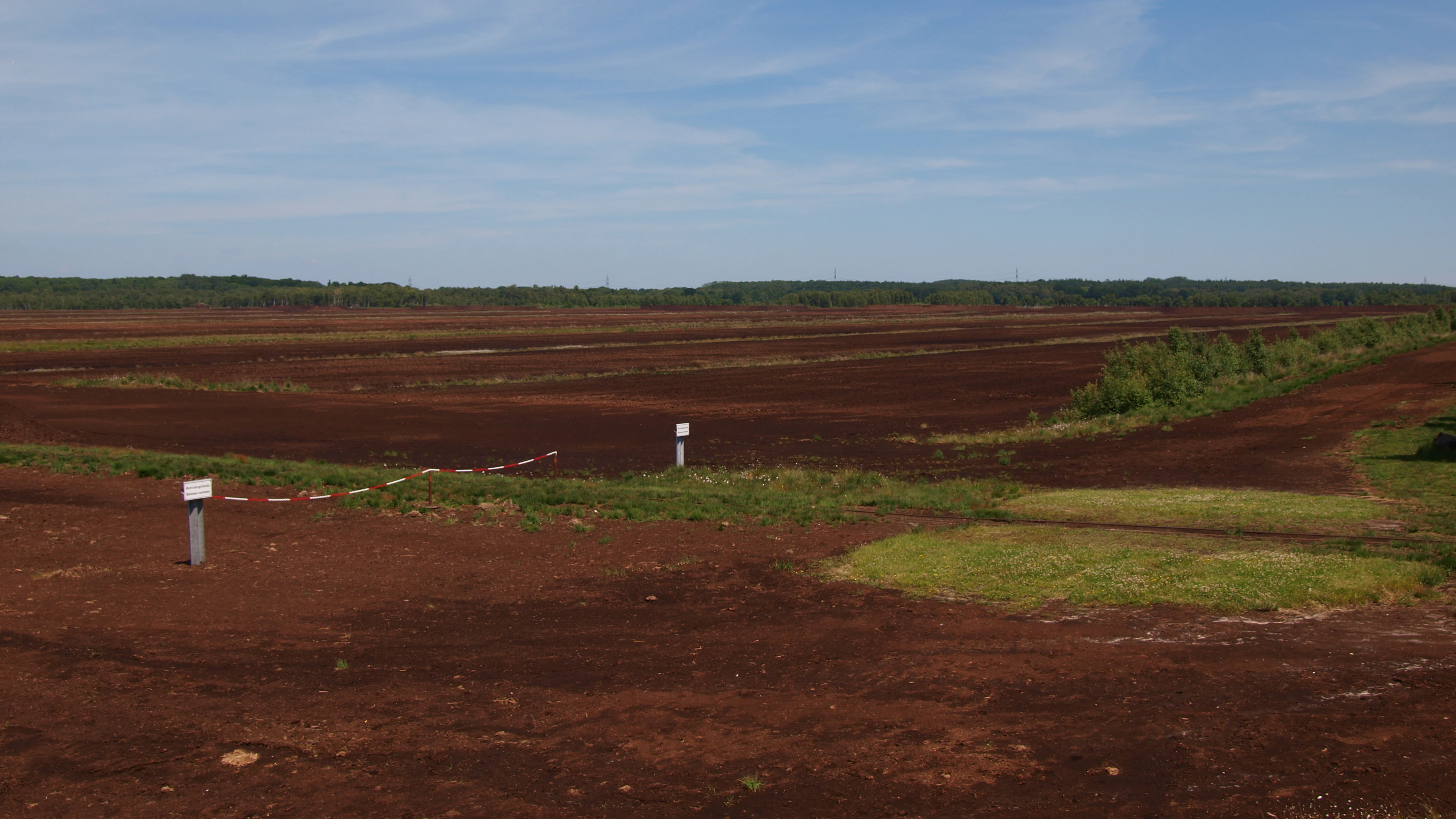 Wide landscape view of a flat, brown, peat extraction field in Himmelmoor under a clear blue sky, with sparse patches of green vegetation and small white markers in the foreground.