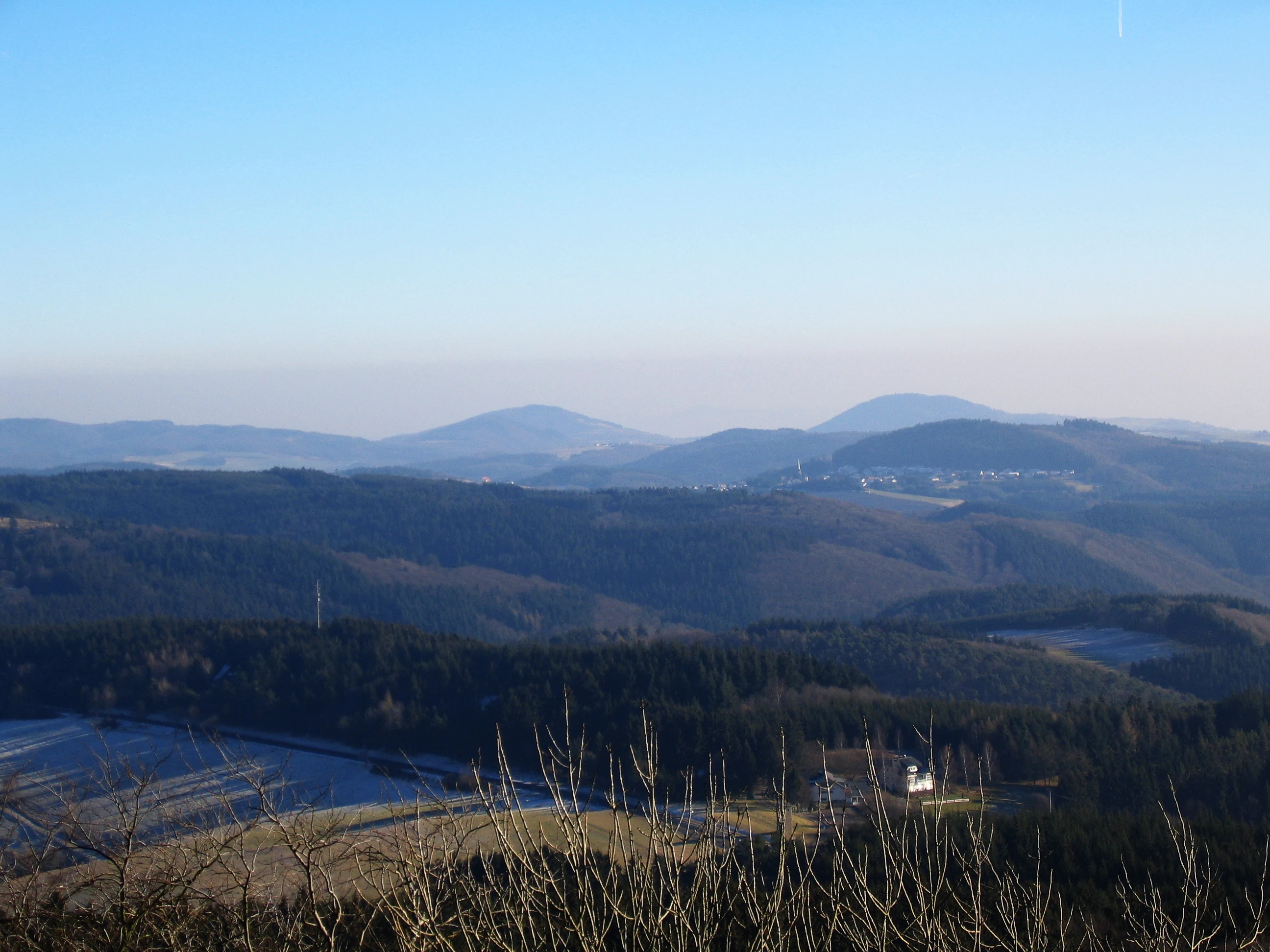 A wide view of rolling forested hills and volcanic peaks under a clear blue sky, seen from a high vantage point.