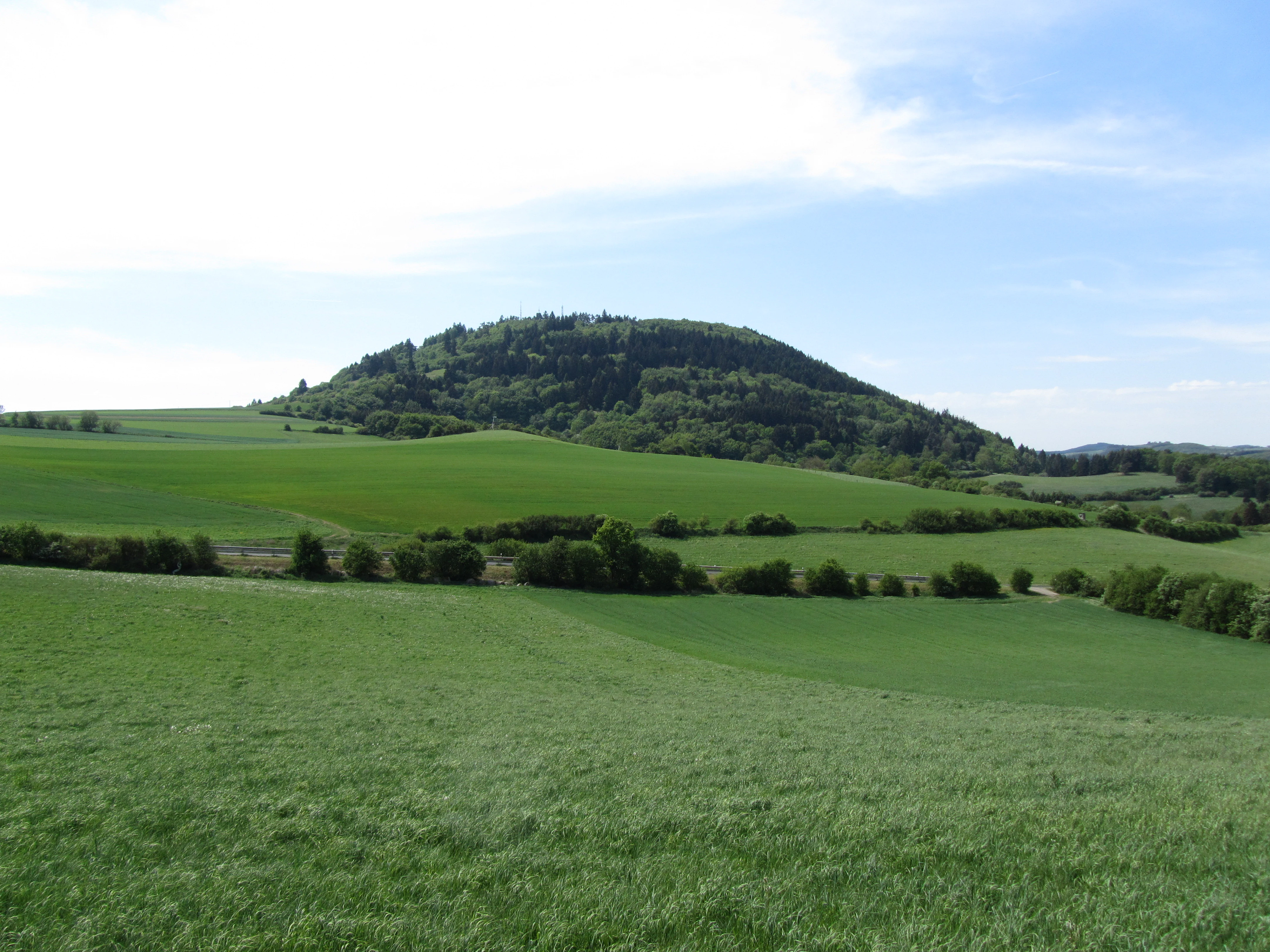 A wide shot of a green, rounded volcanic mountain covered in forest rising above open agricultural fields and rolling meadows under a blue sky.