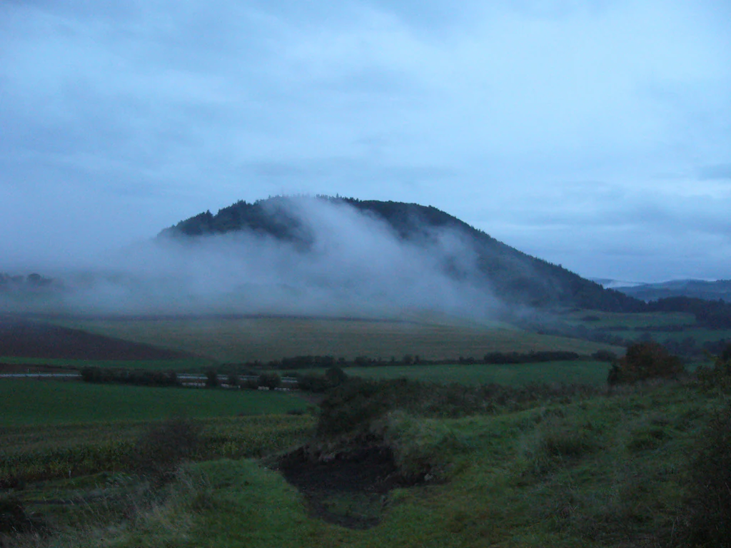 A distant, rounded, forest-covered hill or volcanic cone rising above rolling green fields, with a thick layer of fog wrapping around its midsection under a cloudy, overcast sky.