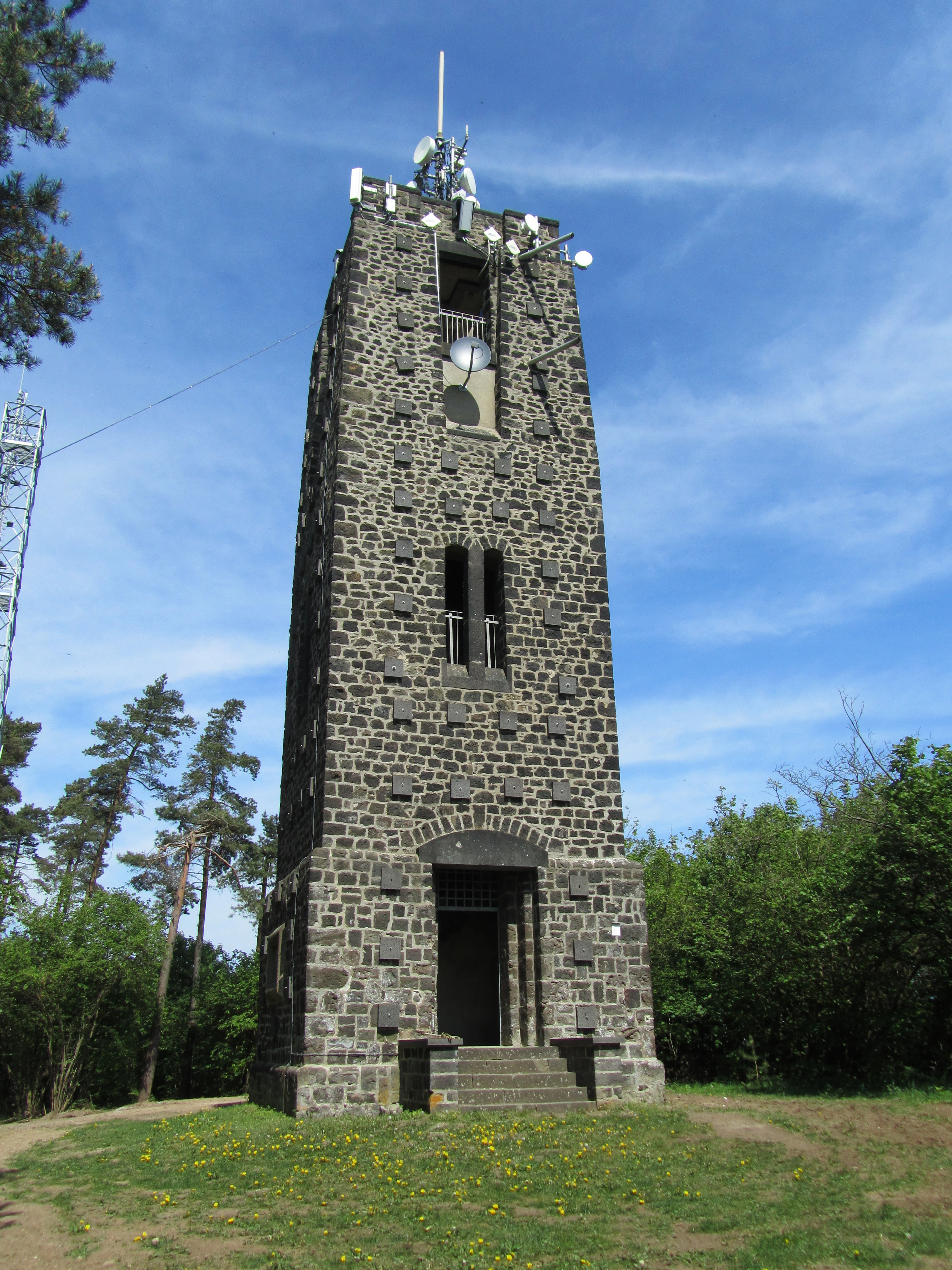 A tall, square observation tower constructed from dark volcanic stone stands in a forest clearing under a bright blue sky.