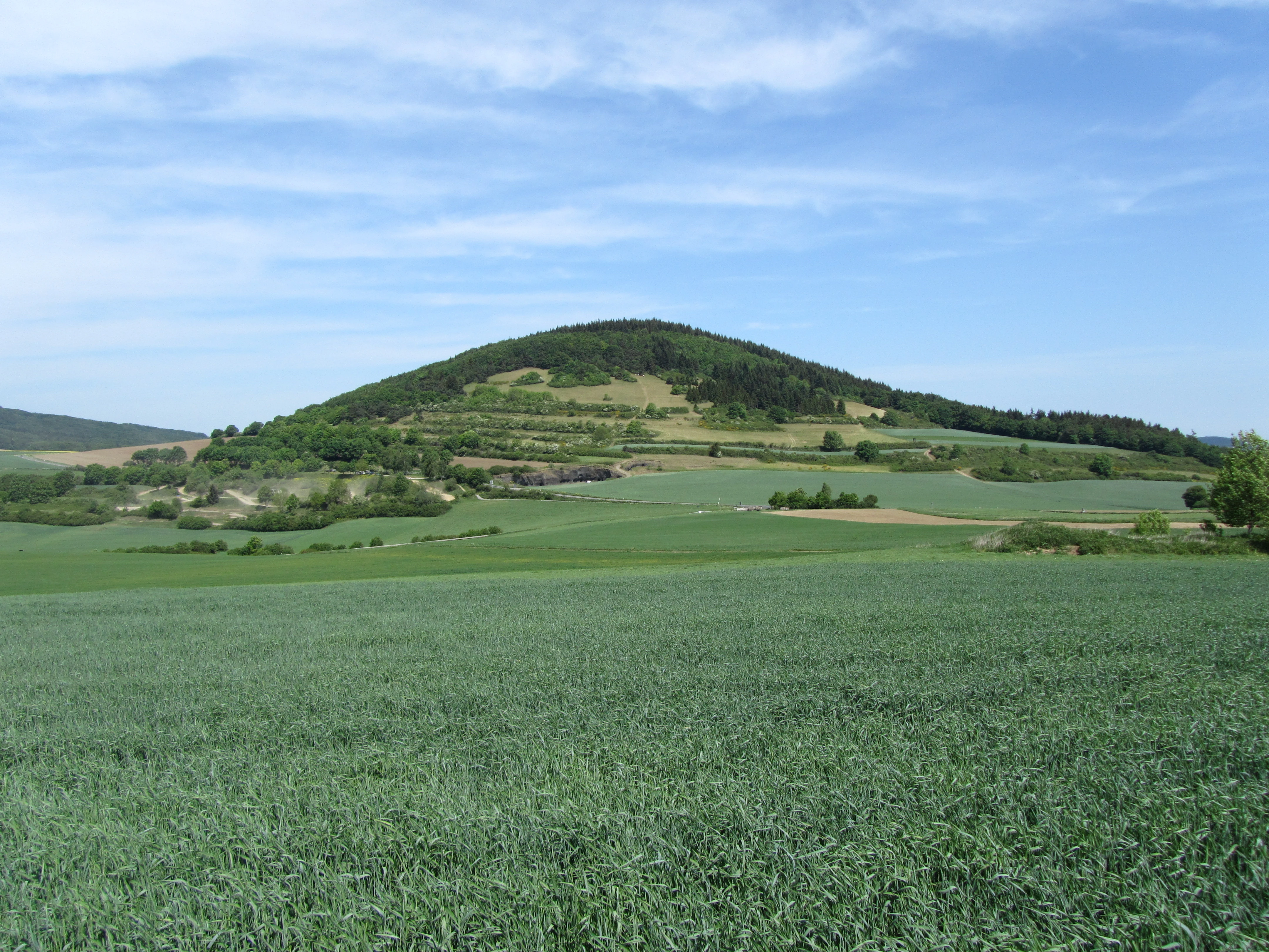 A wide landscape shot of a green volcanic hill, the Hochstein, rising above agricultural fields under a blue sky.