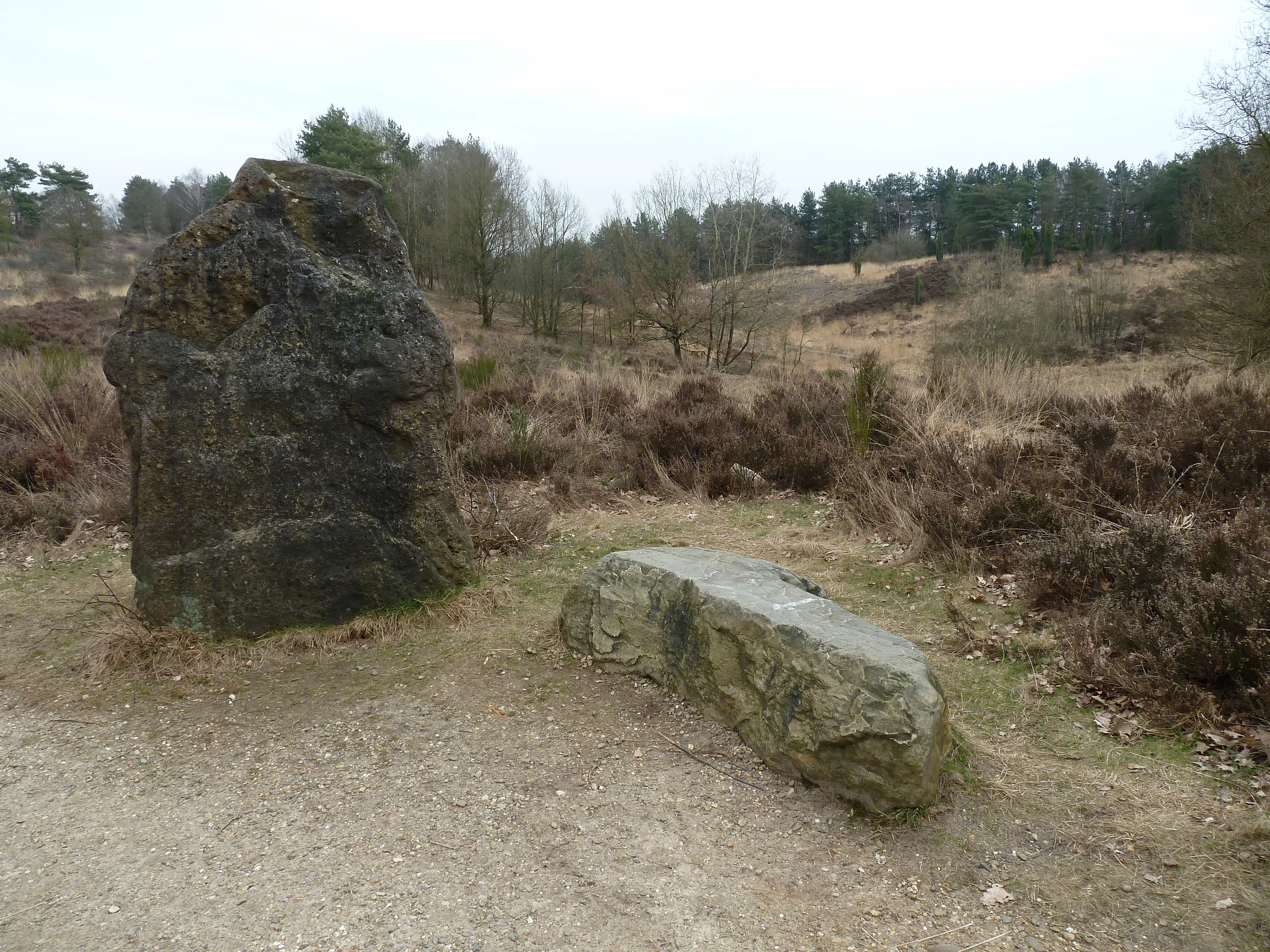 A large standing stone and smaller stone on a gravel path in an open heathland area with dry vegetation and distant trees.