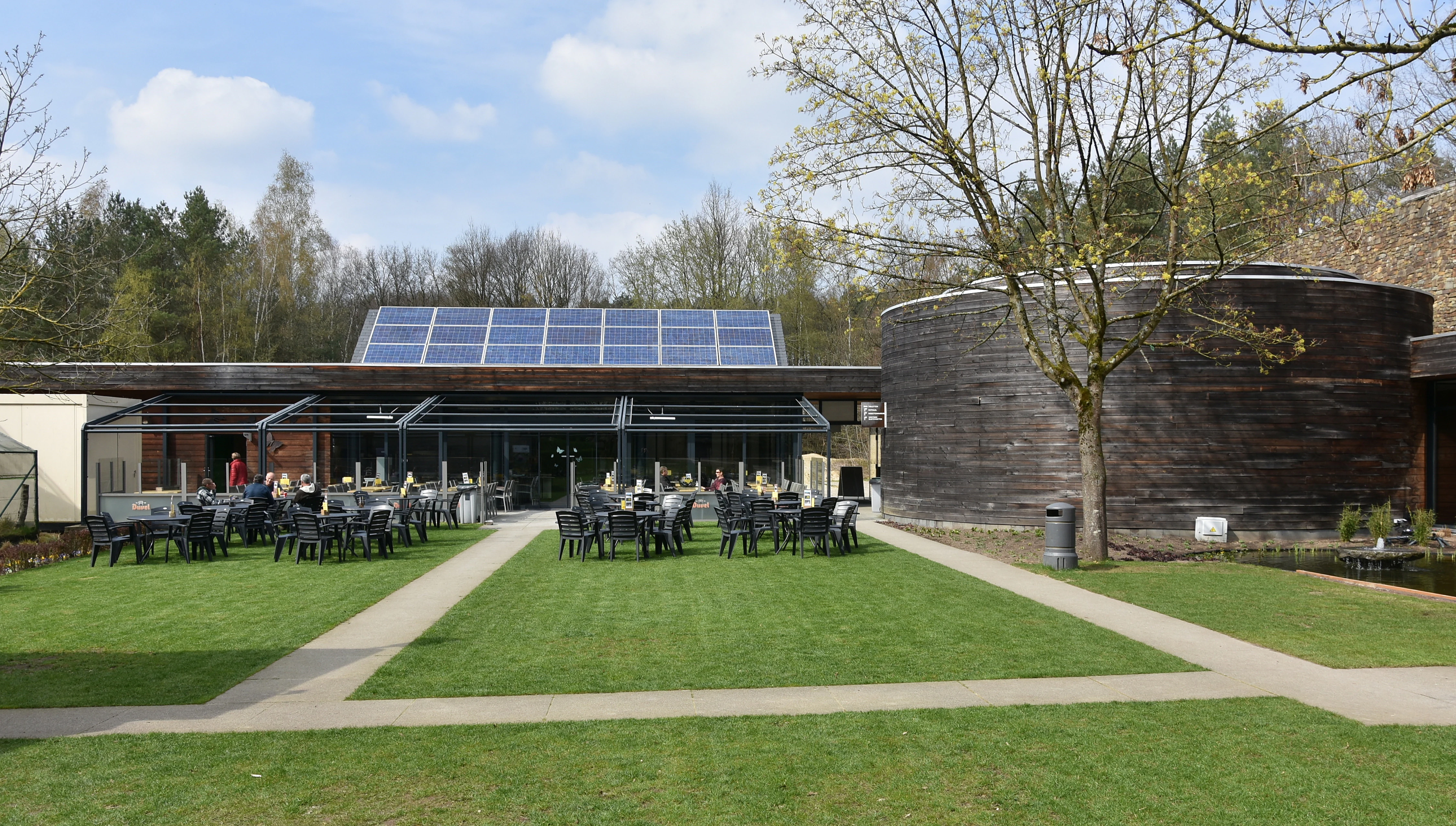 Modern visitor center building with solar panels on roof, outdoor seating area with tables and chairs, and landscaped lawn surrounded by trees and a cylindrical structure