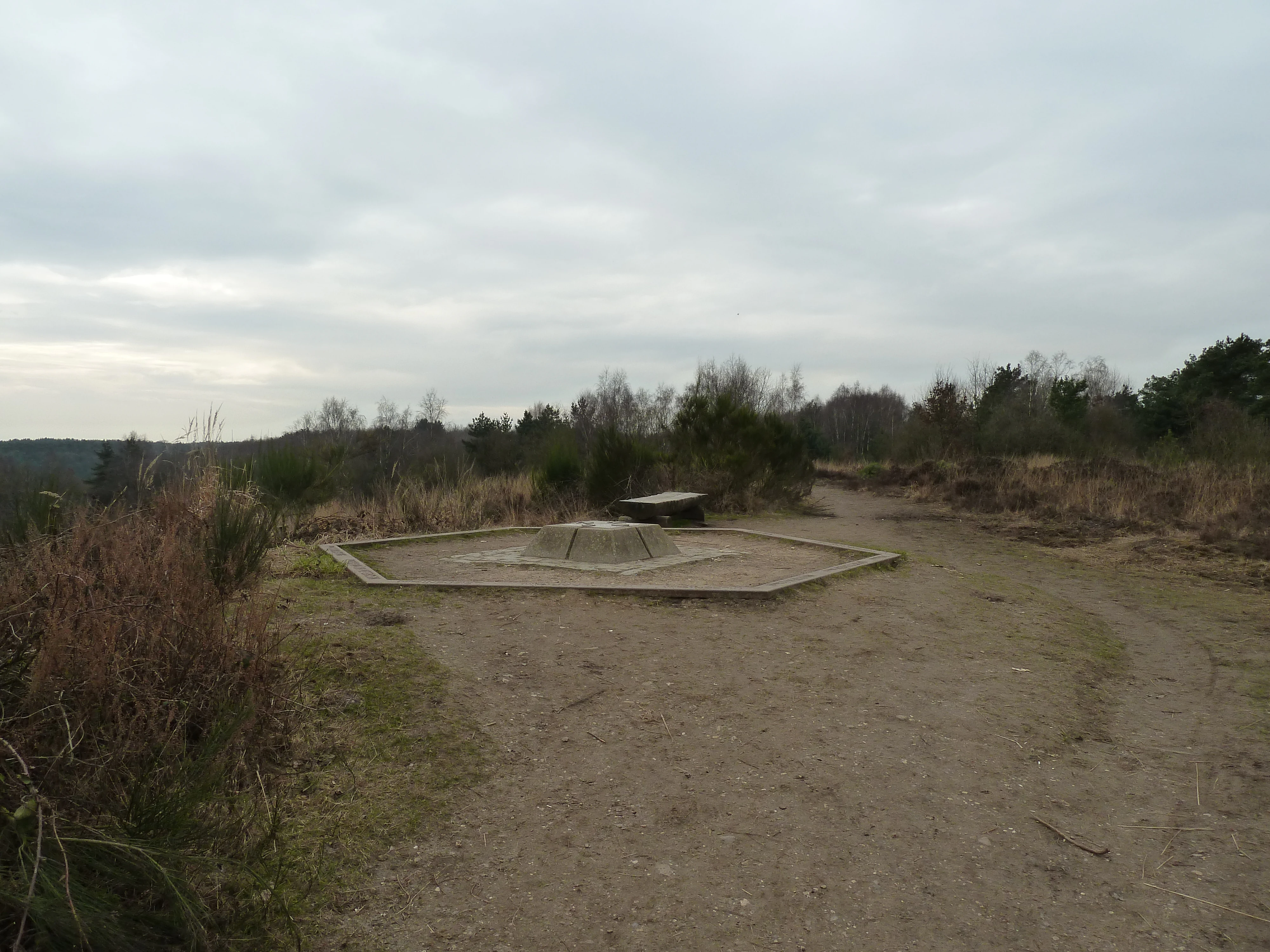Gravel path leading to a hexagonal concrete platform with a small structure, surrounded by dry grass and sparse trees under an overcast sky