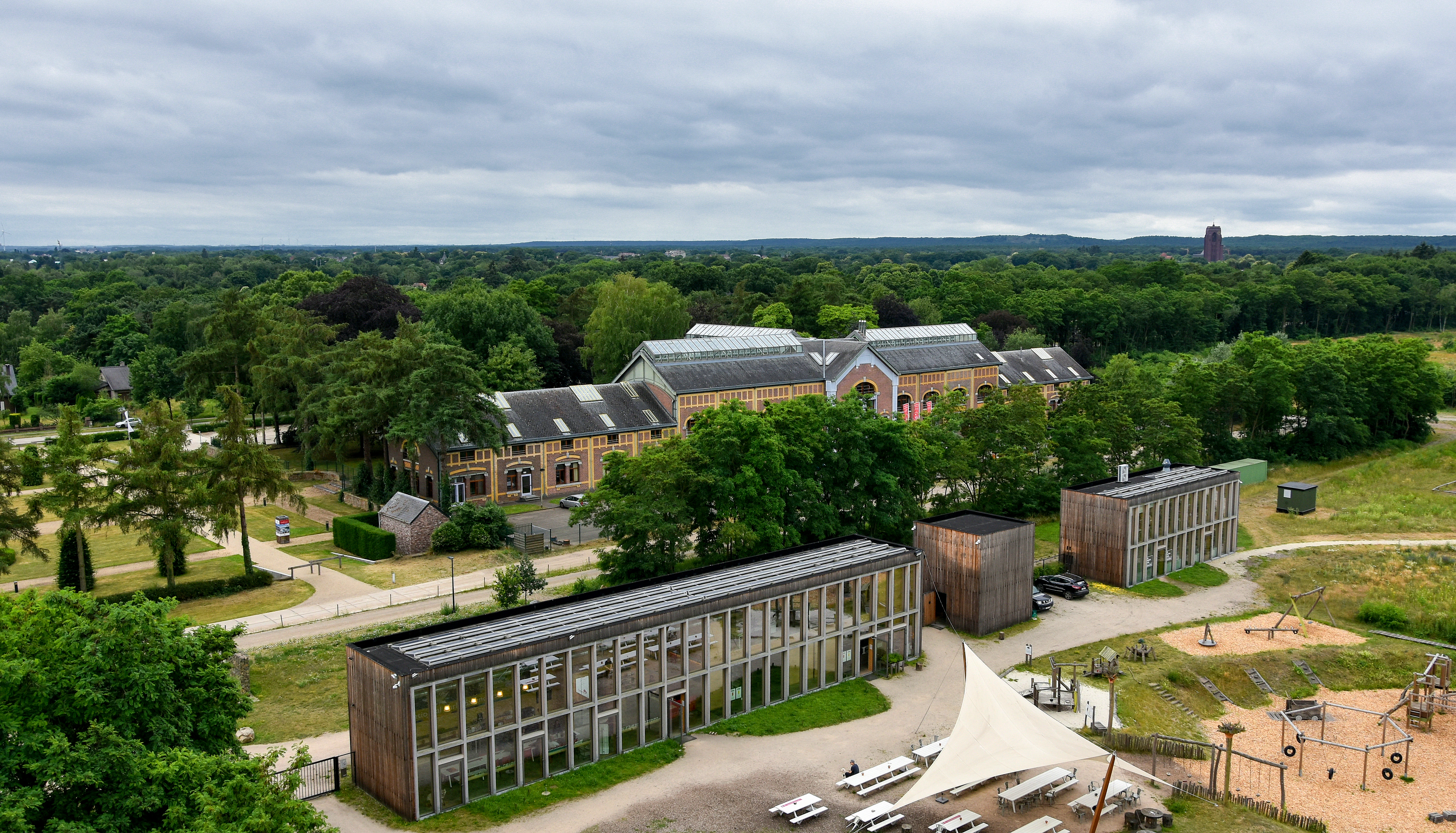 Aerial view of a park entrance complex with multiple buildings including a brick building, glass structures, and a large white tent, surrounded by trees and green fields