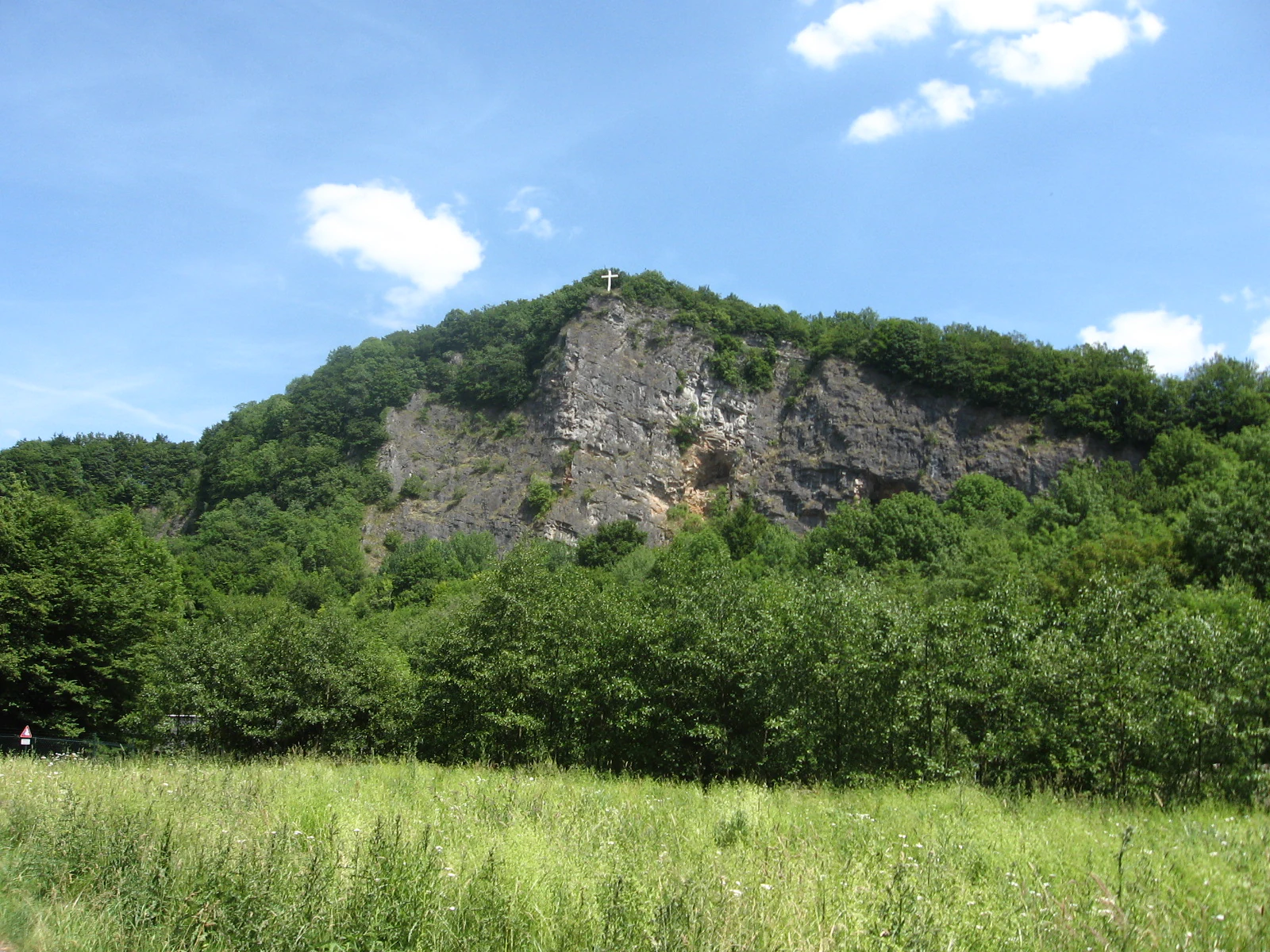 A green, grassy meadow in the foreground leads to a dense forest and a prominent, steep limestone quarry wall with a small cross on its summit against a bright blue sky.