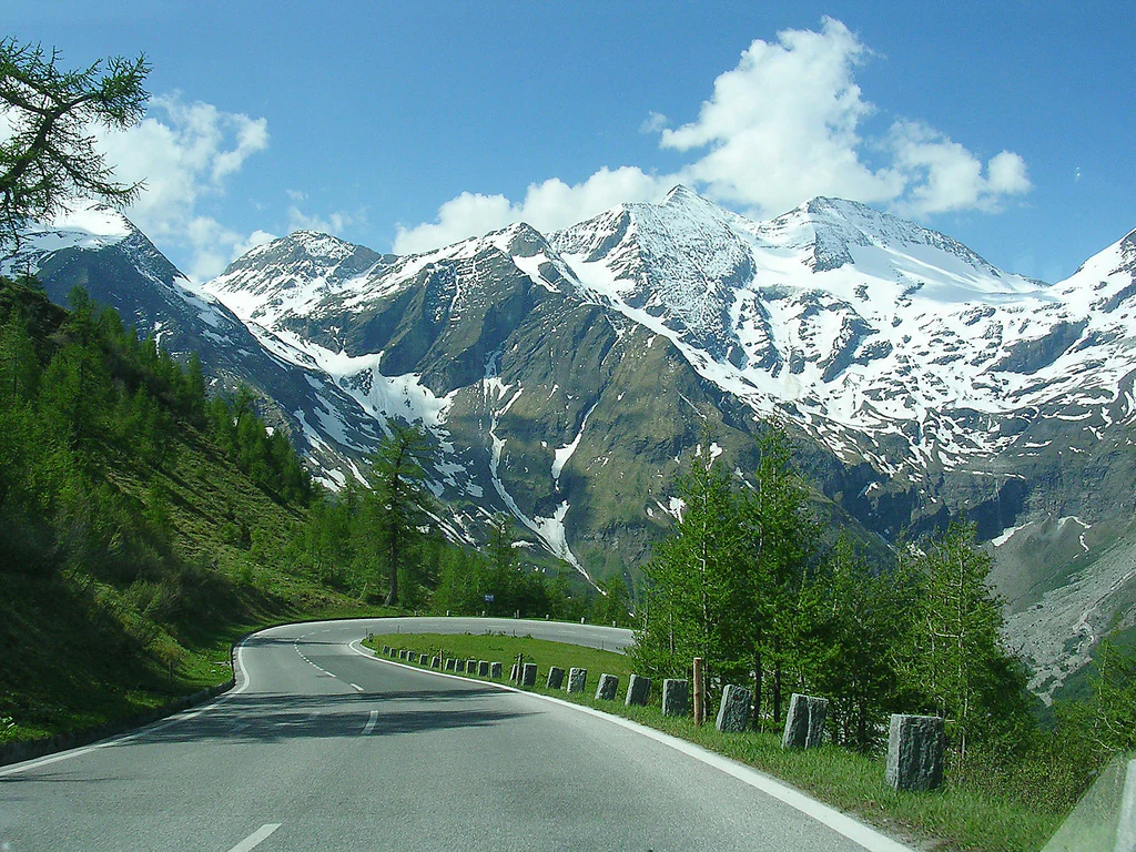 A paved road curves through a mountainous landscape with snow-covered peaks, green trees, and guardrails on a clear day