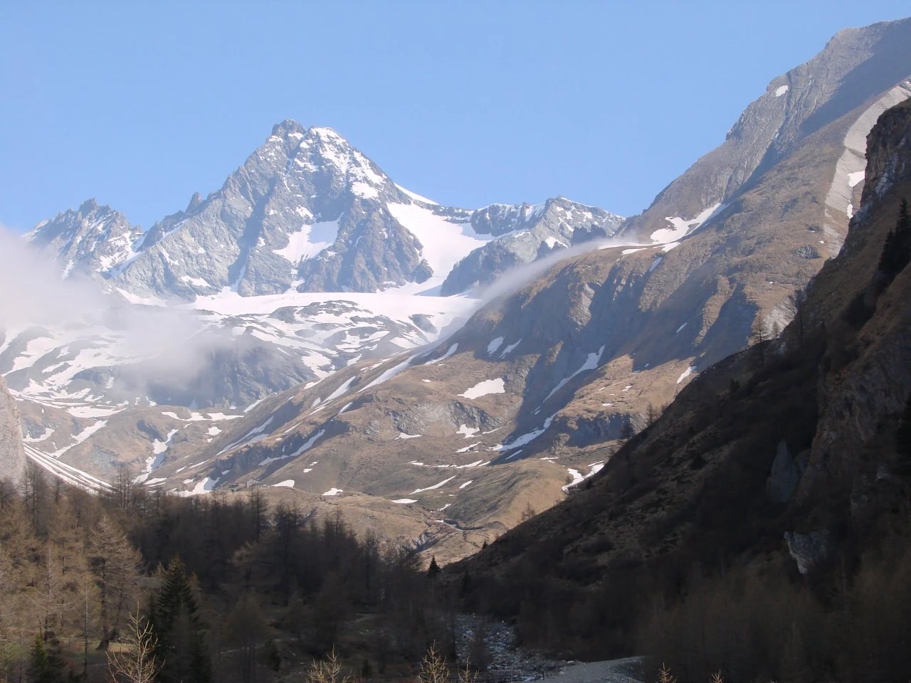 Snow-capped mountain peak with surrounding alpine valleys, forested slopes, and clear blue sky