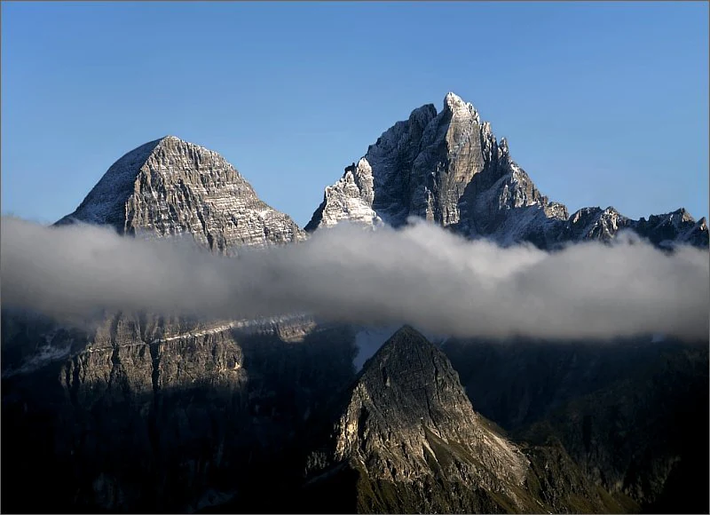 Three mountain peaks with snow on top and rocky slopes, partially obscured by a cloud layer