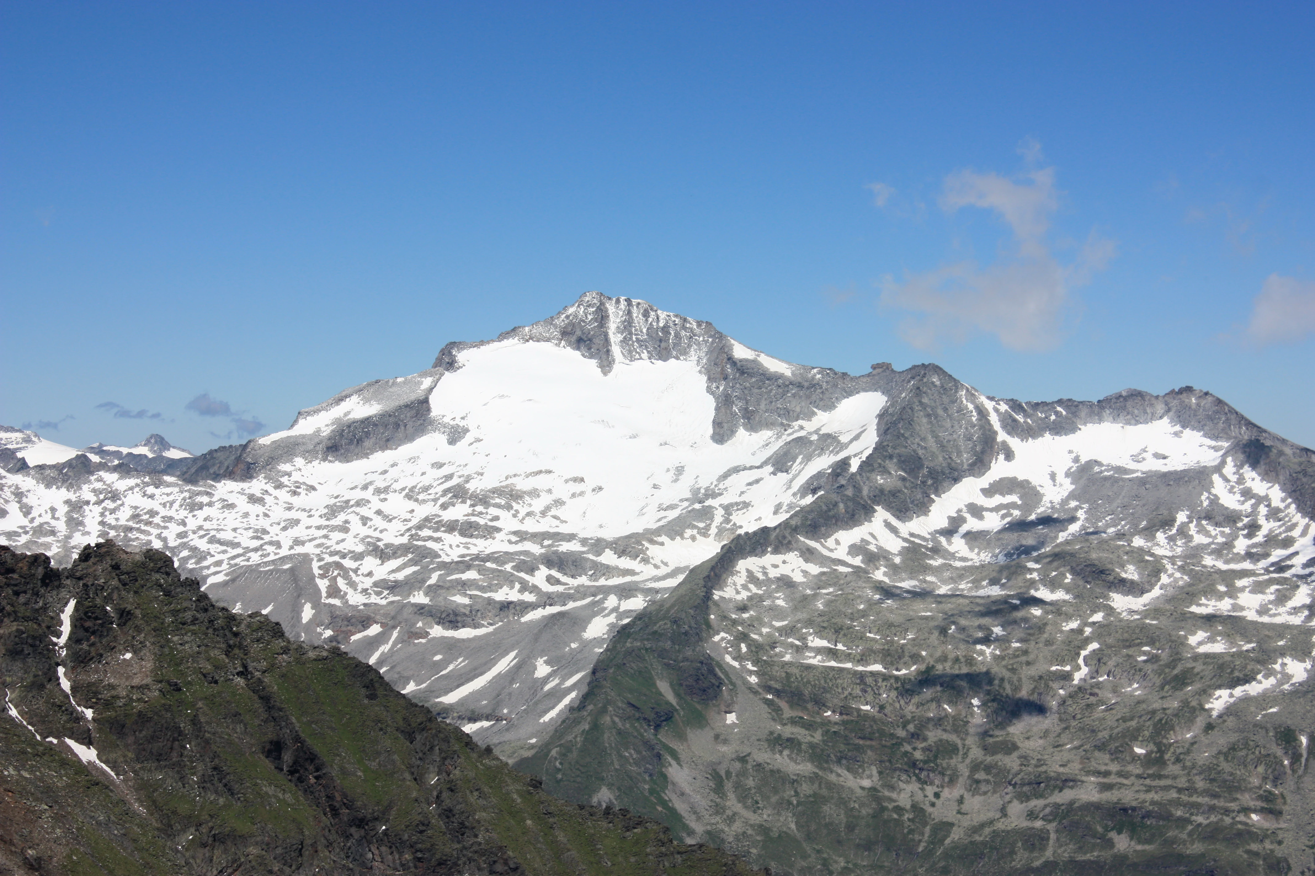Snow-covered mountain peak with rocky slopes and green vegetation under a clear blue sky