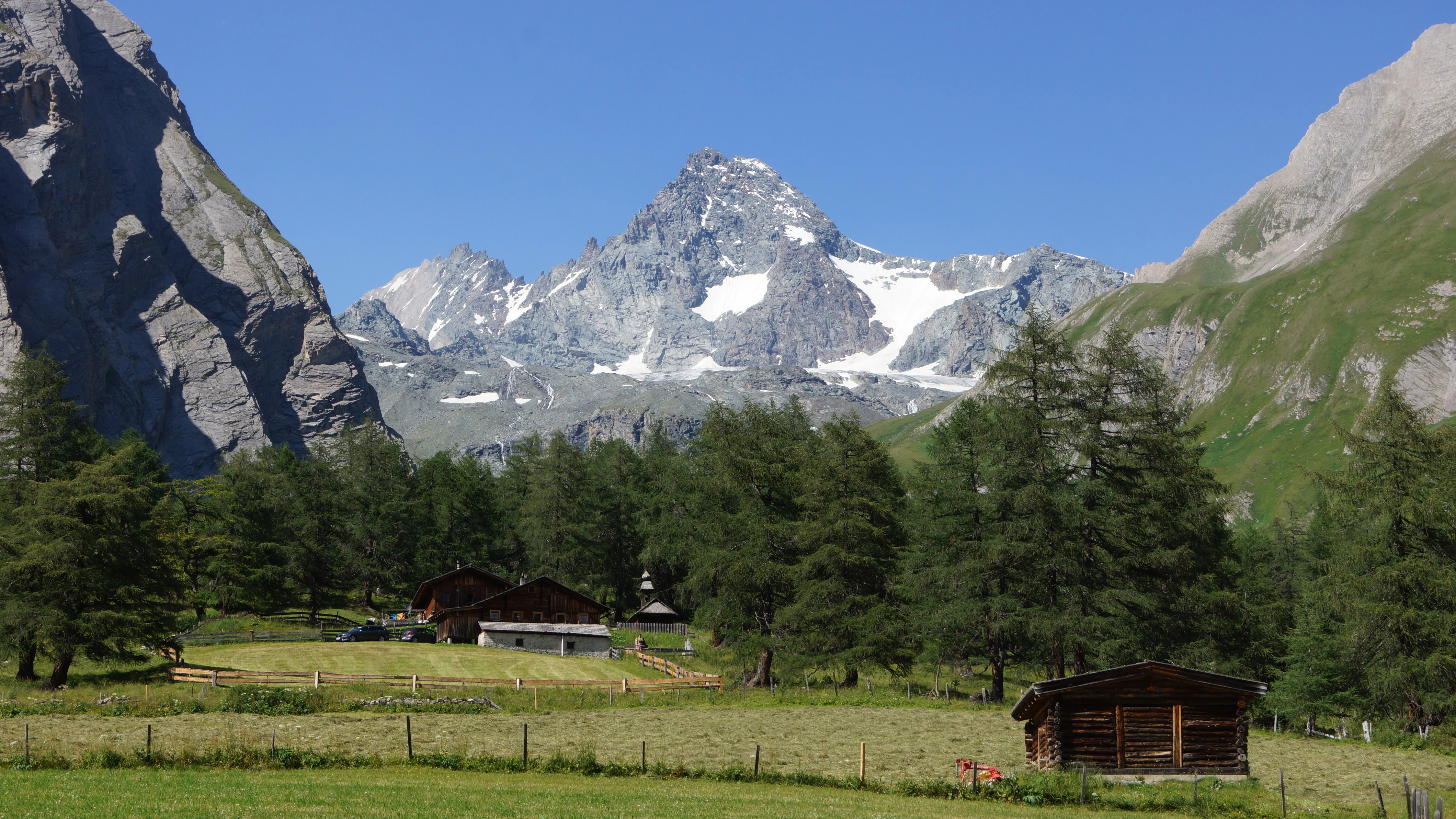 Alpine landscape with mountain valley, wooden huts, and snow-capped mountain peak under clear blue sky