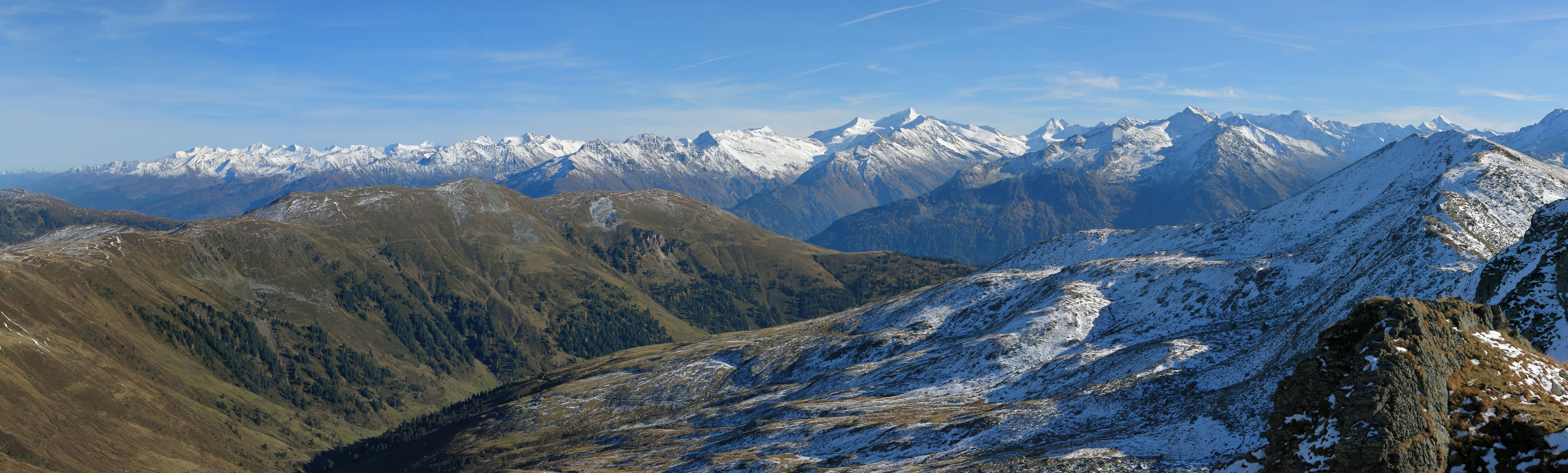 Wide landscape of snow-capped mountains, valleys, and rocky slopes under a clear blue sky