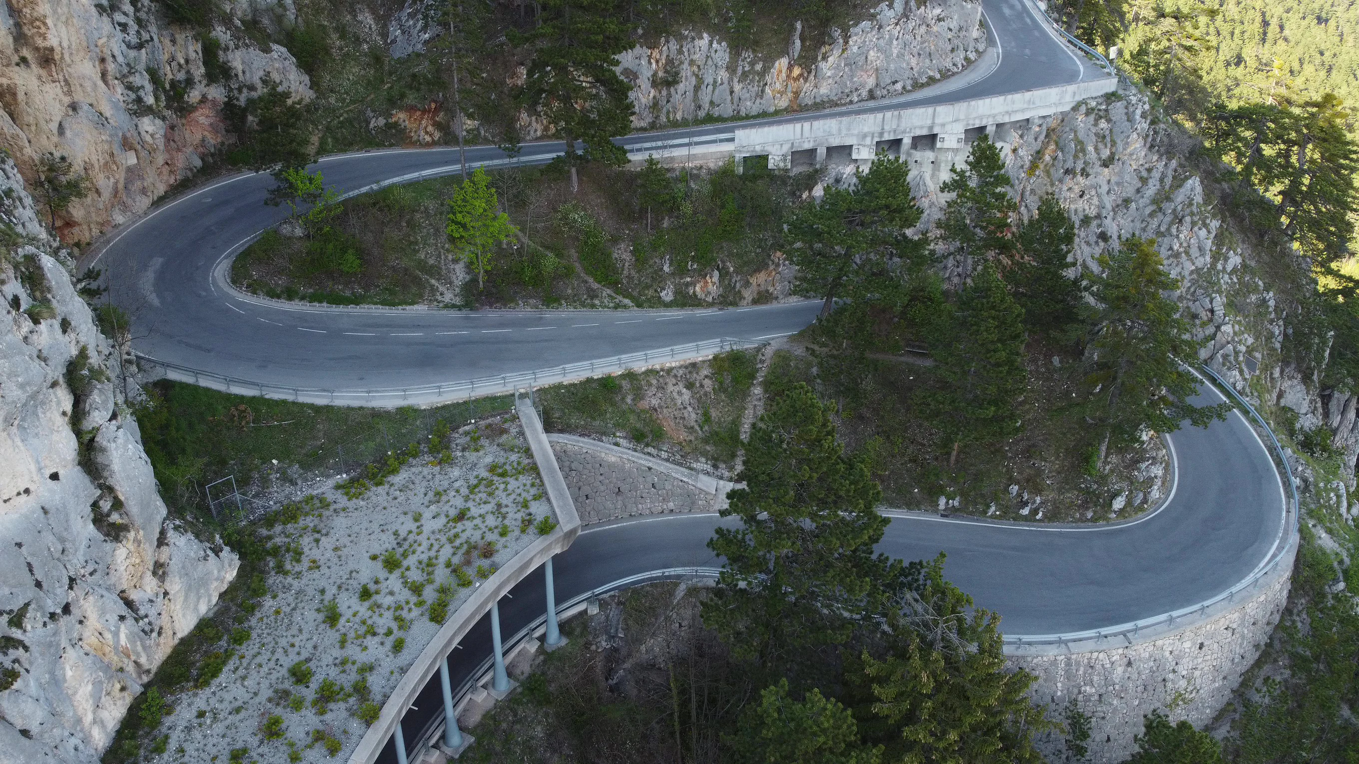 An aerial view of a narrow, winding road with multiple hairpin turns carved into steep, pale limestone mountain cliffs, with sparse trees along the roadside.