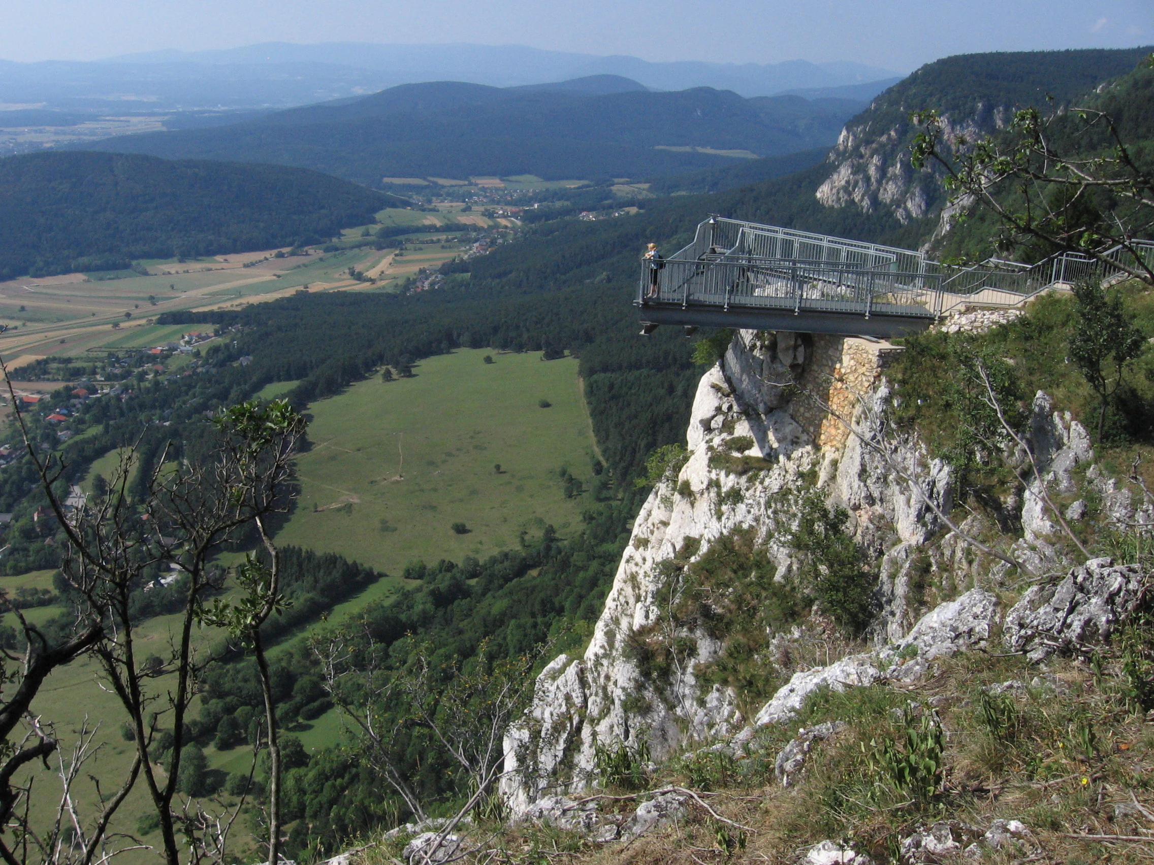 A cantilevered metal observation deck extending over a steep limestone cliff with a wide view of the valley below.