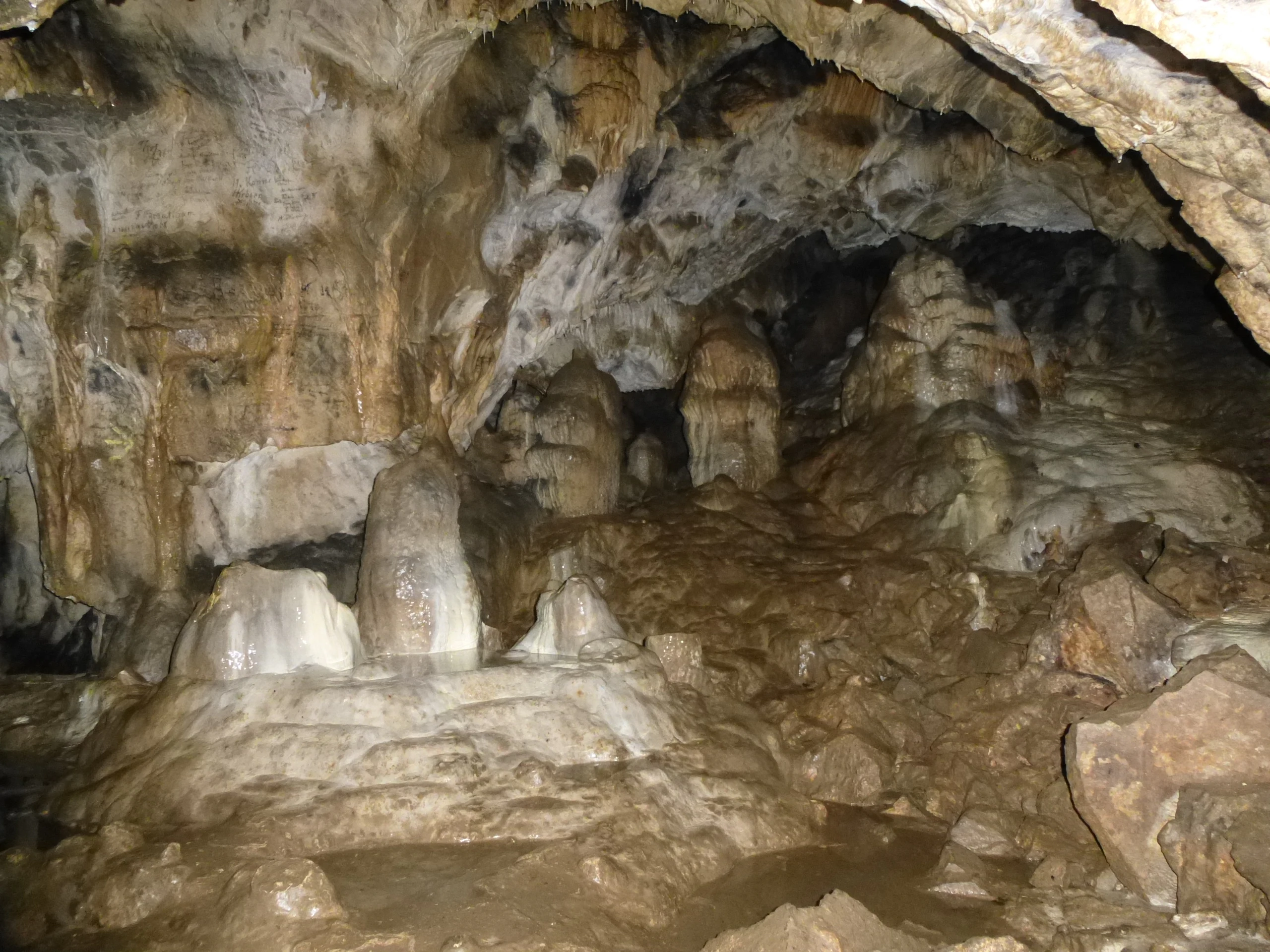 Interior view of a cave chamber with large stalagmites and rocky formations on the floor and ceiling.