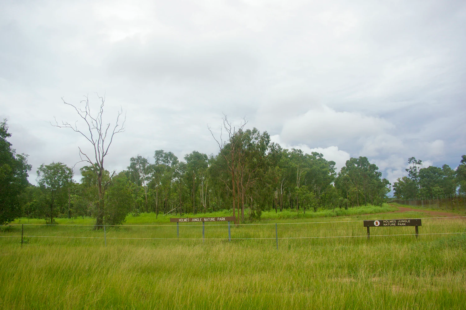 A grassy field leads to a line of trees with park identification signs visible in the middle ground under a cloudy sky.