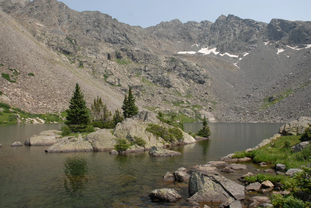 A calm, clear lake with a rocky island featuring several small evergreen trees in the center, surrounded by steep, craggy mountain slopes containing patches of snow.