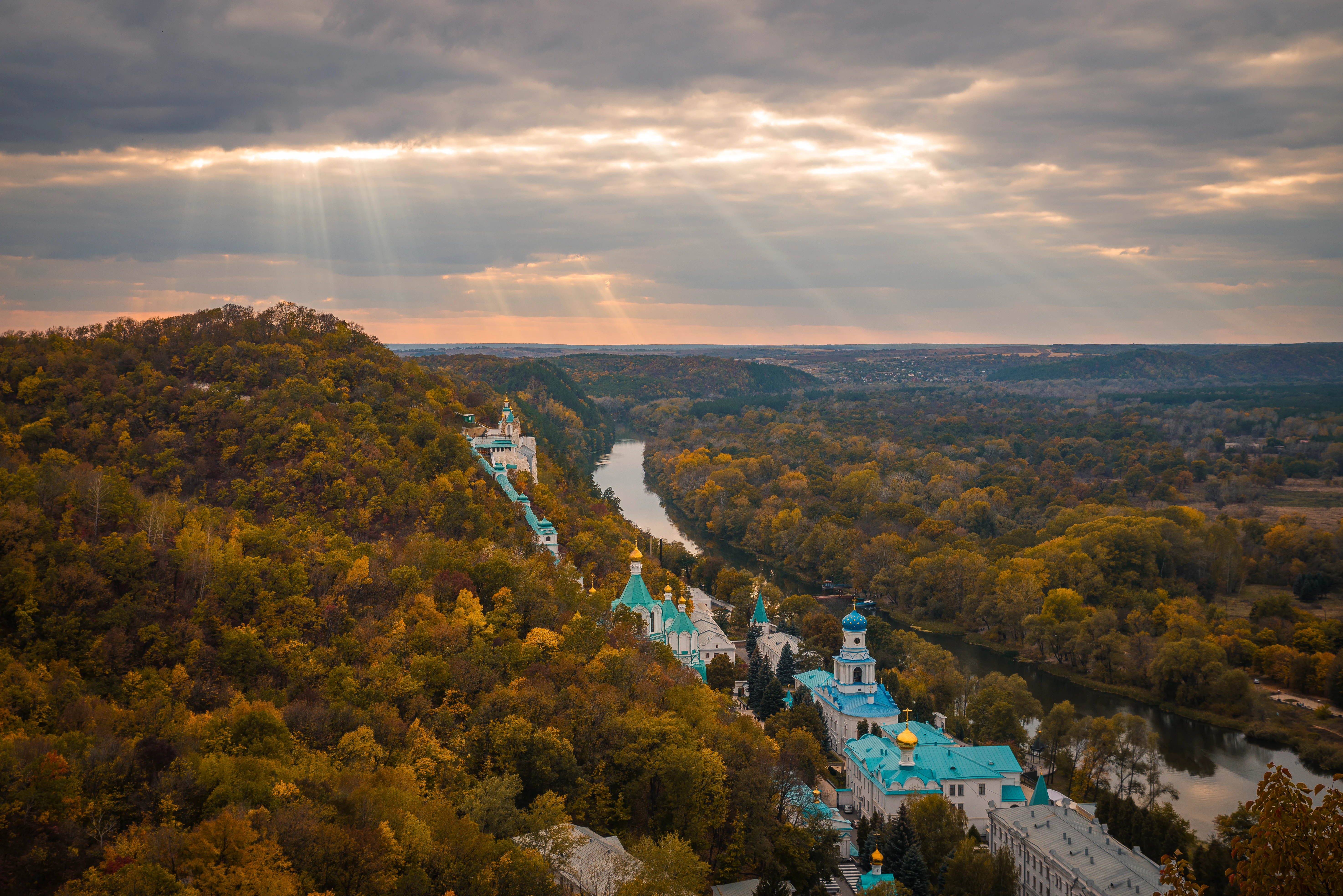 Aerial view of the Sviatohirsk Lavra monastery complex with blue domes situated on a forested hillside above the Donets River under a cloudy sky with sunbeams