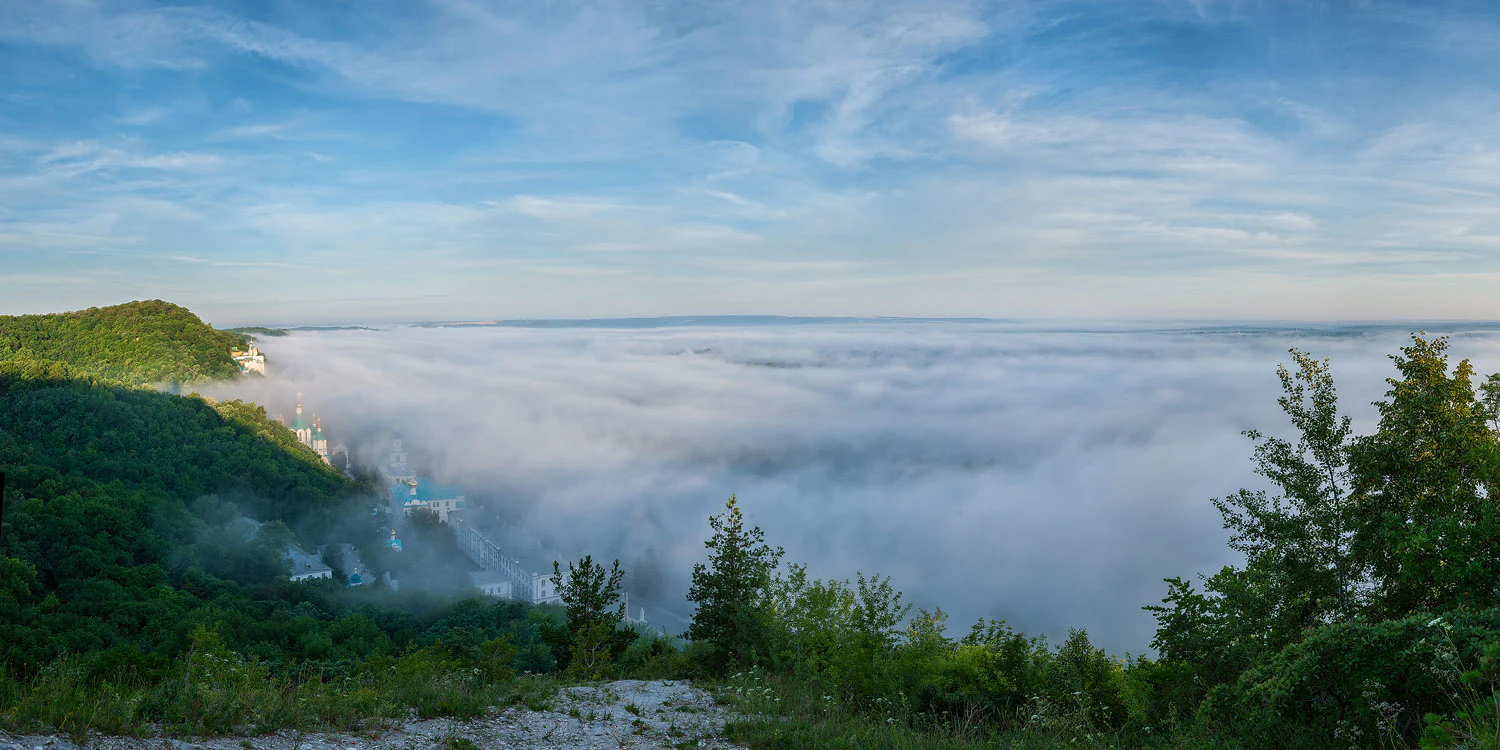 Panoramic landscape view featuring a fog-filled valley, forested hills on the left, and trees in the foreground under a partly cloudy sky