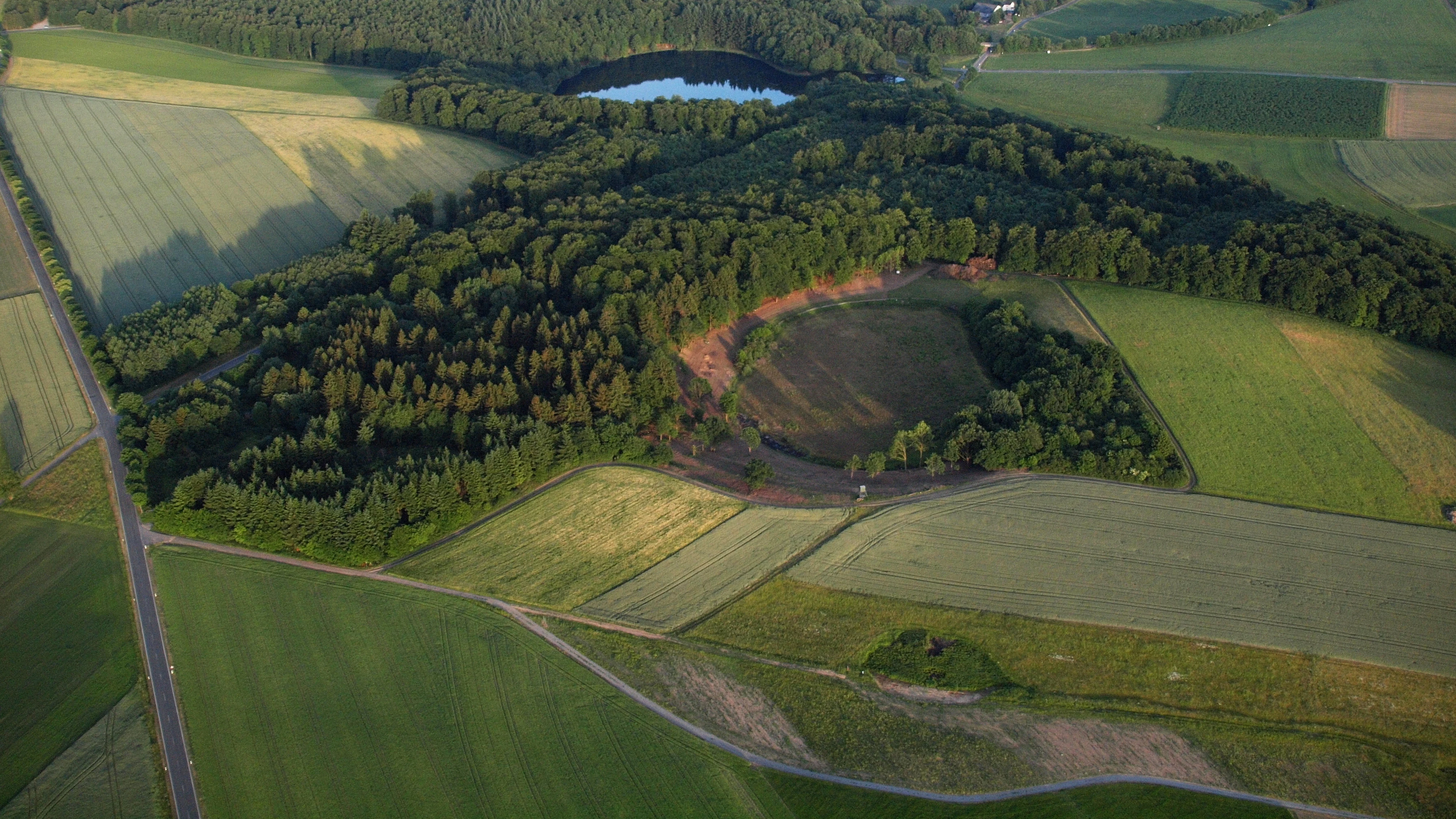 An aerial view of lush green landscapes showing three distinct volcanic maar craters, including Holzmaar lake, surrounded by fields and patches of forest.
