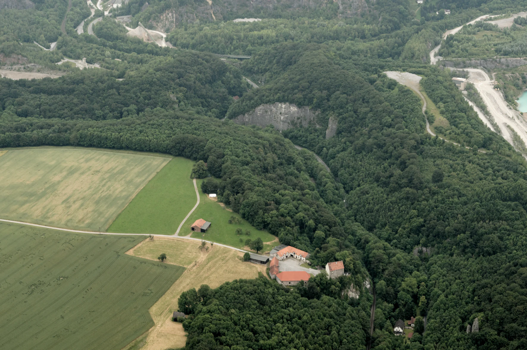 Una fotografía aérea que muestra un valle verde y exuberante con bosques densos, campos agrícolas, un castillo con tejado rojo y una cantera de piedra caliza al fondo.