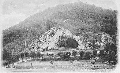 Fotografía histórica en blanco y negro de 1901 que muestra la gran entrada de una cueva en un acantilado de piedra caliza, rodeada por colinas densamente boscosas con un campo de hierba y árboles dispersos en primer plano