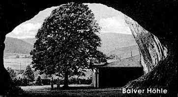 Fotografía histórica en blanco y negro de 1902 que muestra una vista desde el interior de la cueva de Balve, donde la entrada de la cueva enmarca un gran árbol de hoja caduca, un pequeño edificio y colinas onduladas al fondo. El texto Balver Hohle aparece en la esquina inferior derecha.