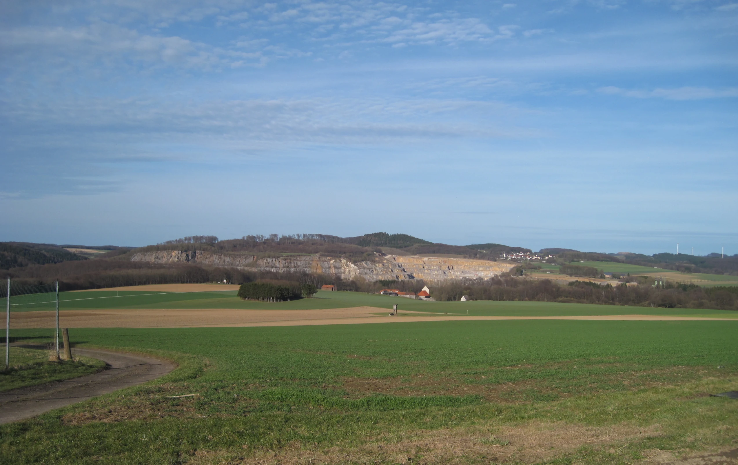 Una vista panorámica de campos agrícolas verdes que conducen a un gran acantilado de piedra caliza de color claro en la distancia, bajo un cielo azul con nubes dispersas.