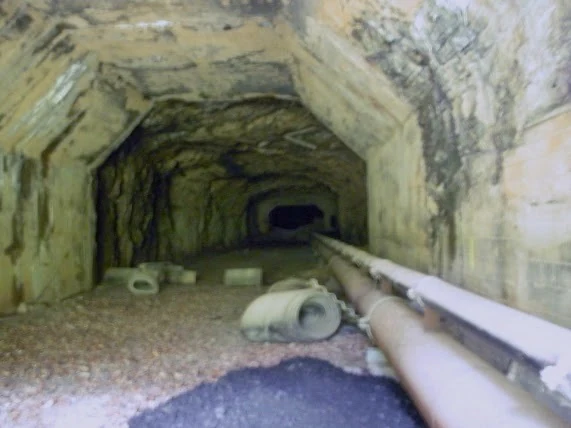 Interior de un túnel rocoso con paredes y techo de piedra rugosa, tuberías de servicio a lo largo del lado derecho, bloques de hormigón y escombros dispersos por el suelo de grava, y una abertura oscura visible al fondo.