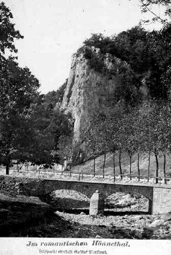 Fotografía histórica en blanco y negro de 1902 que muestra un puente de piedra con arco sobre el lecho rocoso de un río, con un gran acantilado de piedra caliza y árboles que se alzan detrás. El texto en la parte inferior dice 'Im romantischen Hönnetal'.