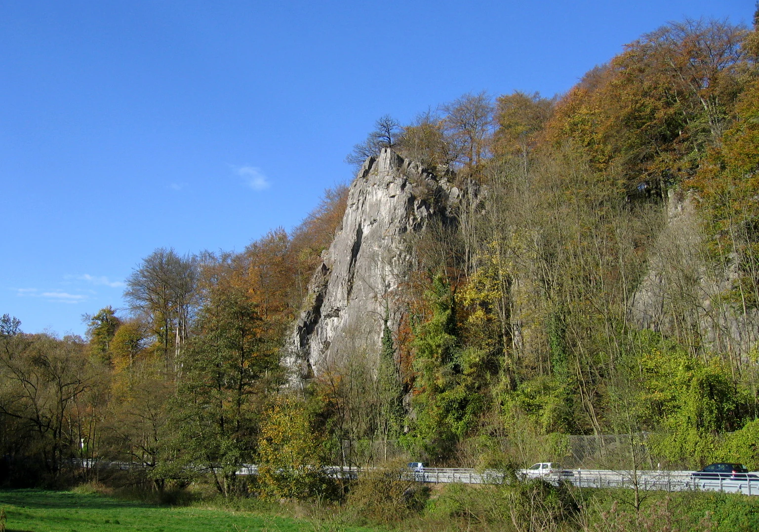 Una alta formación rocosa de piedra caliza rodeada de árboles otoñales, con una carretera y vehículos a su base bajo un cielo azul despejado.