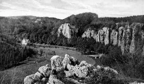 Fotografía histórica en blanco y negro que muestra un valle con prominentes acantilados de piedra caliza, un bosque denso, un río que serpentea por el fondo del valle y algunos edificios a lo lejos.