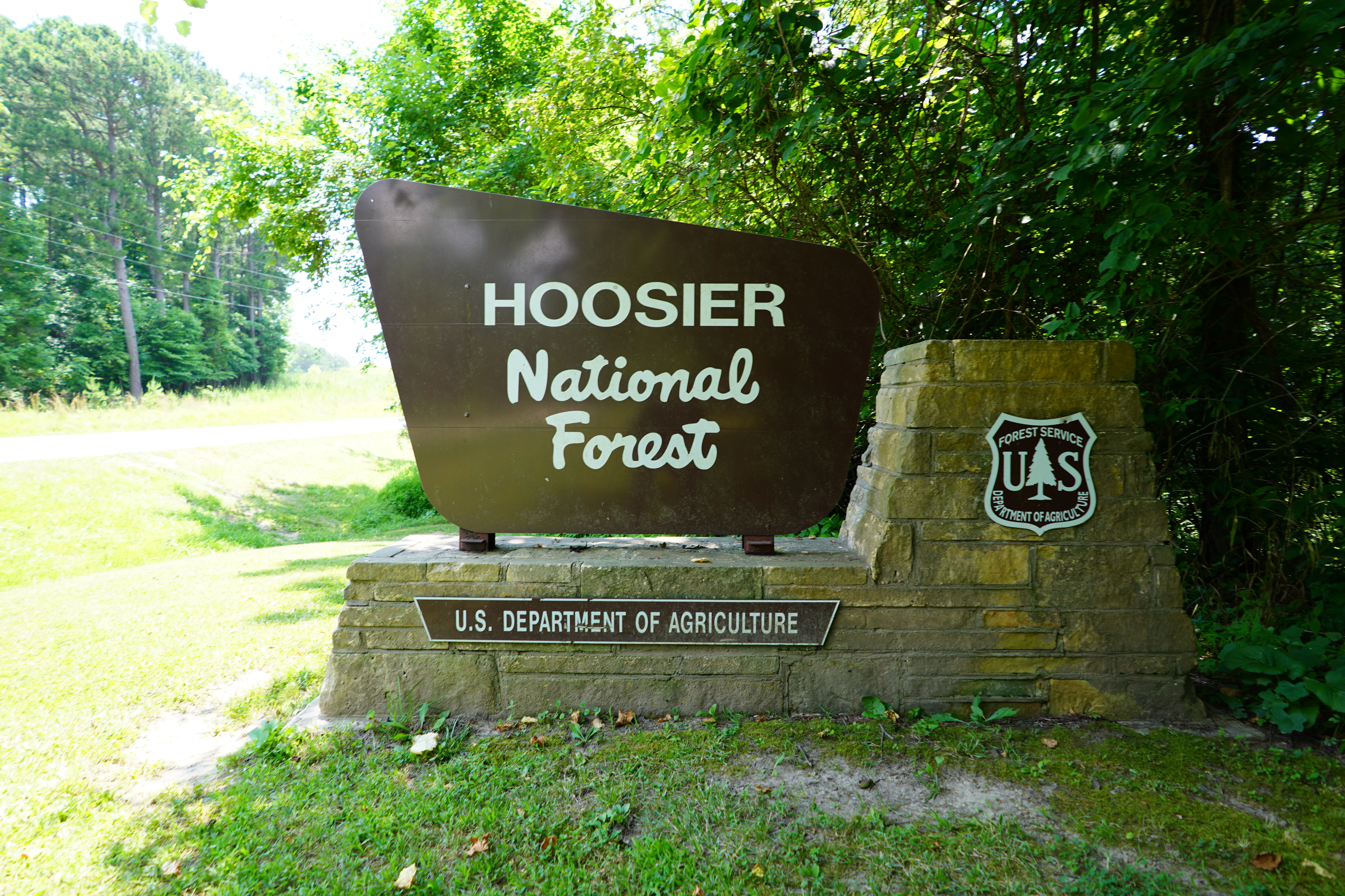 A brown, trapezoidal entrance sign for Hoosier National Forest, mounted on a stone base with the U.S. Forest Service logo to the right, surrounded by lush green trees.