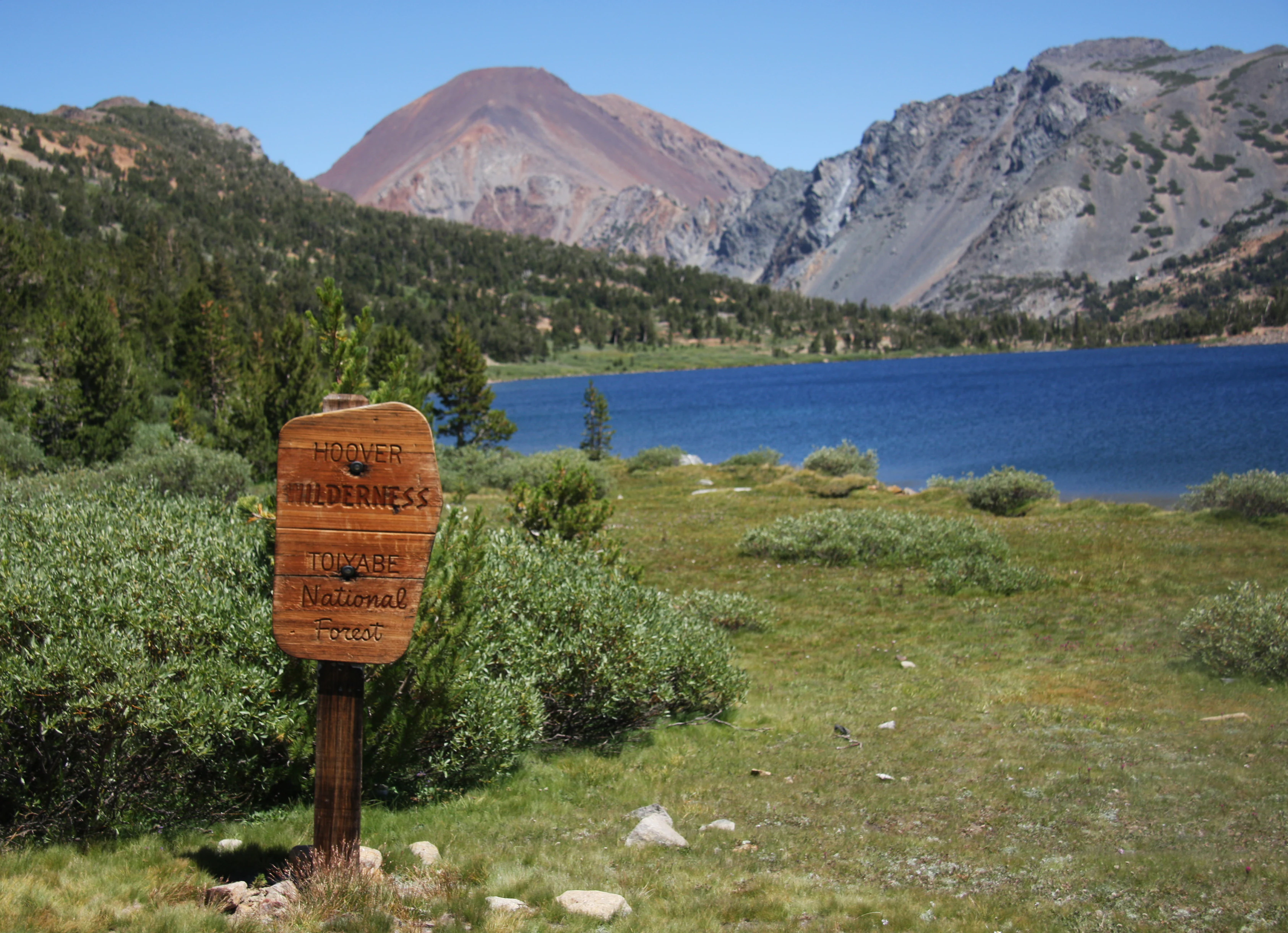 A wooden sign stands in a grassy field with a deep blue lake and rocky mountain peaks in the background.