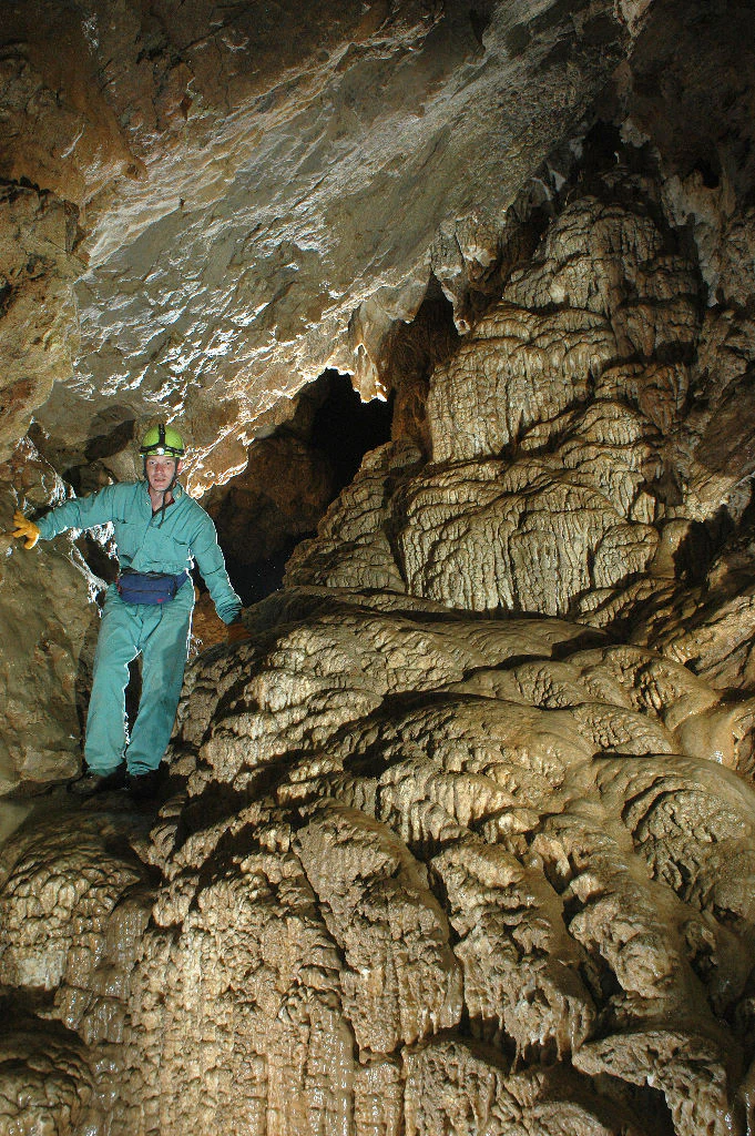 A person wearing a caving suit and helmet explores a limestone cave with flowing rock formations.