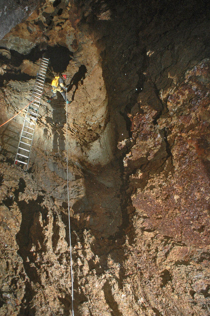 A person wearing a helmet and backpack descends a vertical cave wall inside a cavern, using a metal ladder and a safety rope against dark, rugged rock formations.