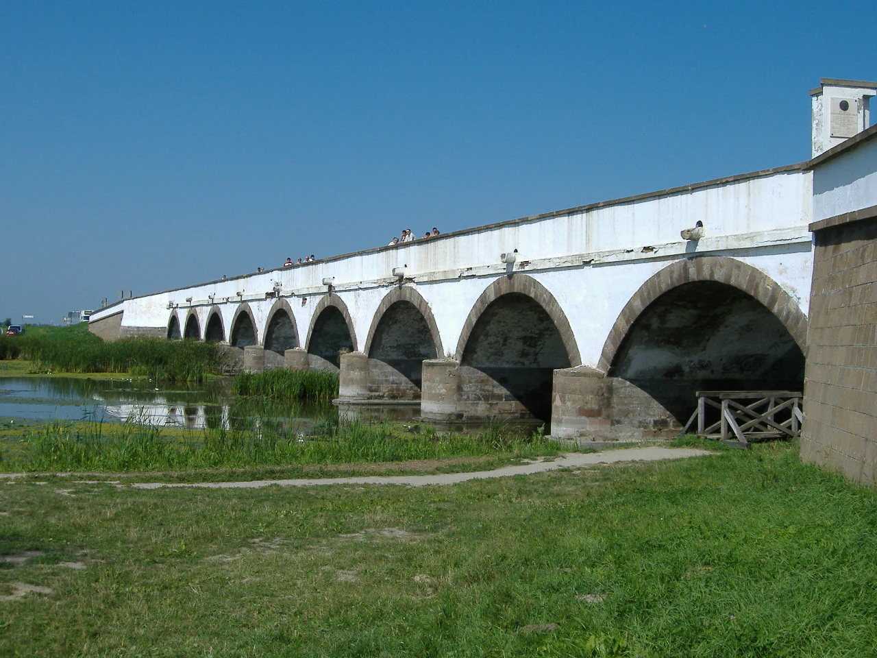 Stone bridge with multiple arches spanning a body of water, surrounded by grassy landscape under clear blue sky