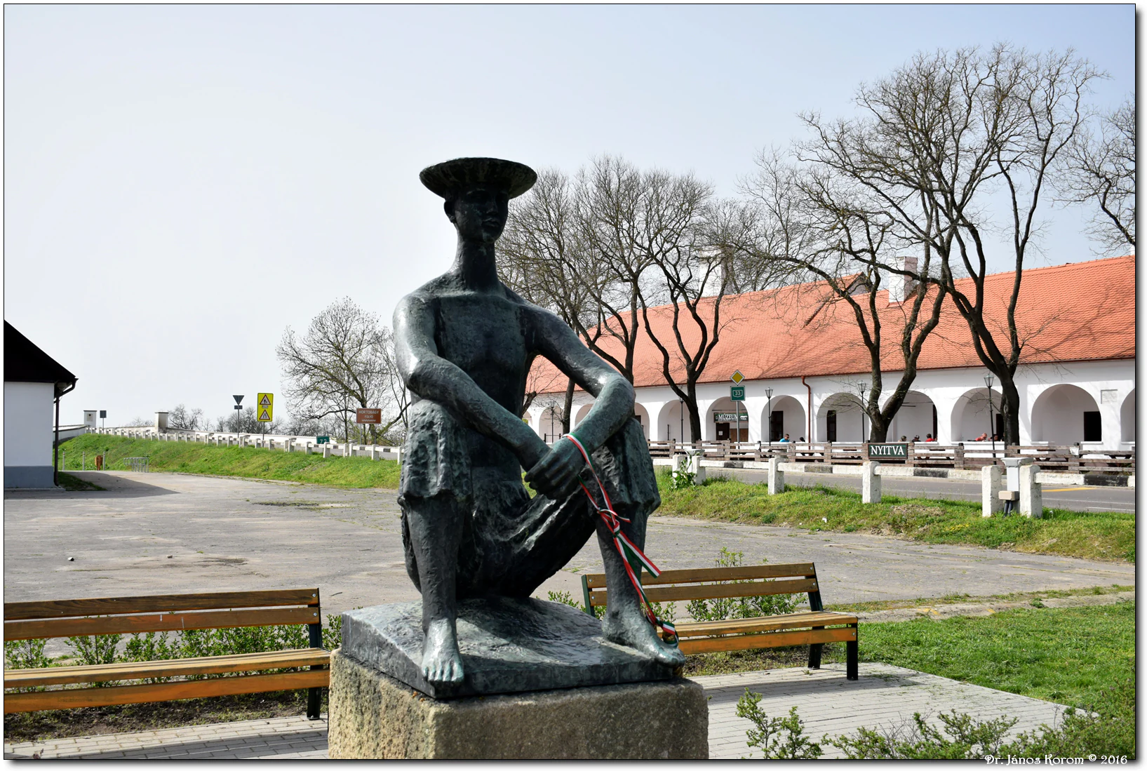 Bronze statue of a seated shepherd boy wearing a hat and holding a rope, with benches and a red-roofed building in the background