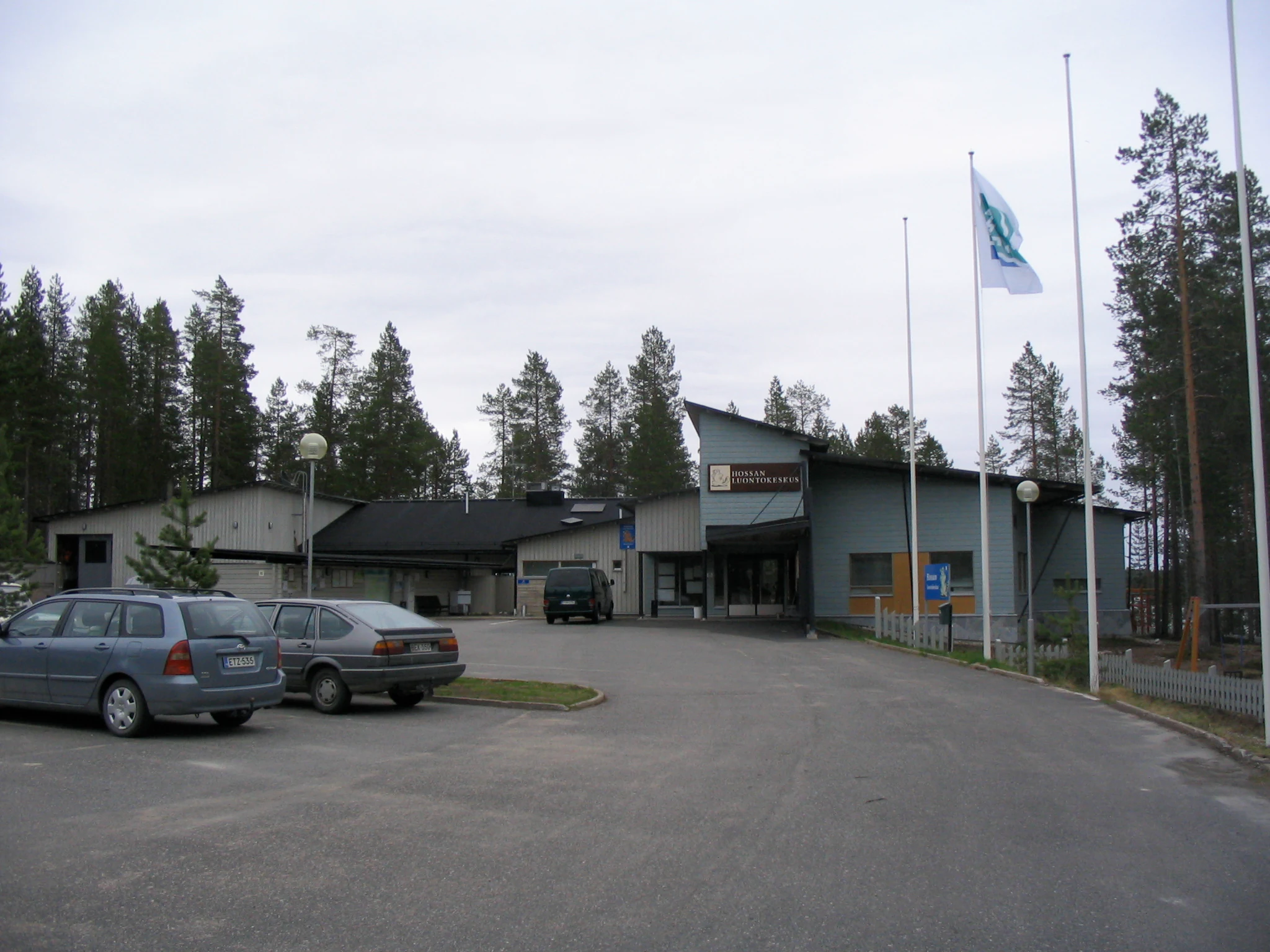 Modern visitor center building with parking lot, cars, and flagpoles surrounded by pine forest