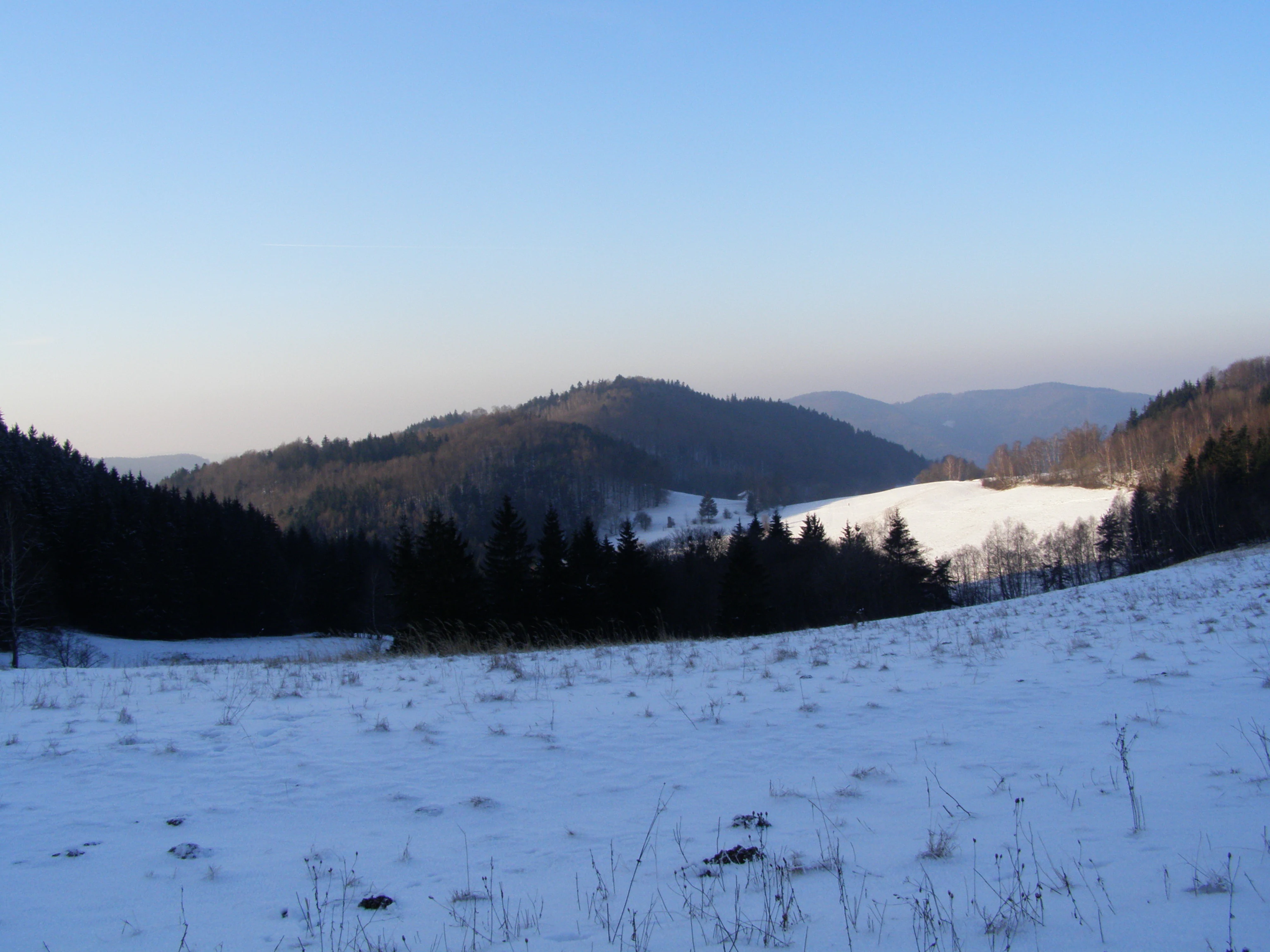 A wide, snowy landscape of the Hostýnské vrchy hills under a clear blue sky, featuring snow-covered fields in the foreground and dark forested hills in the distance.
