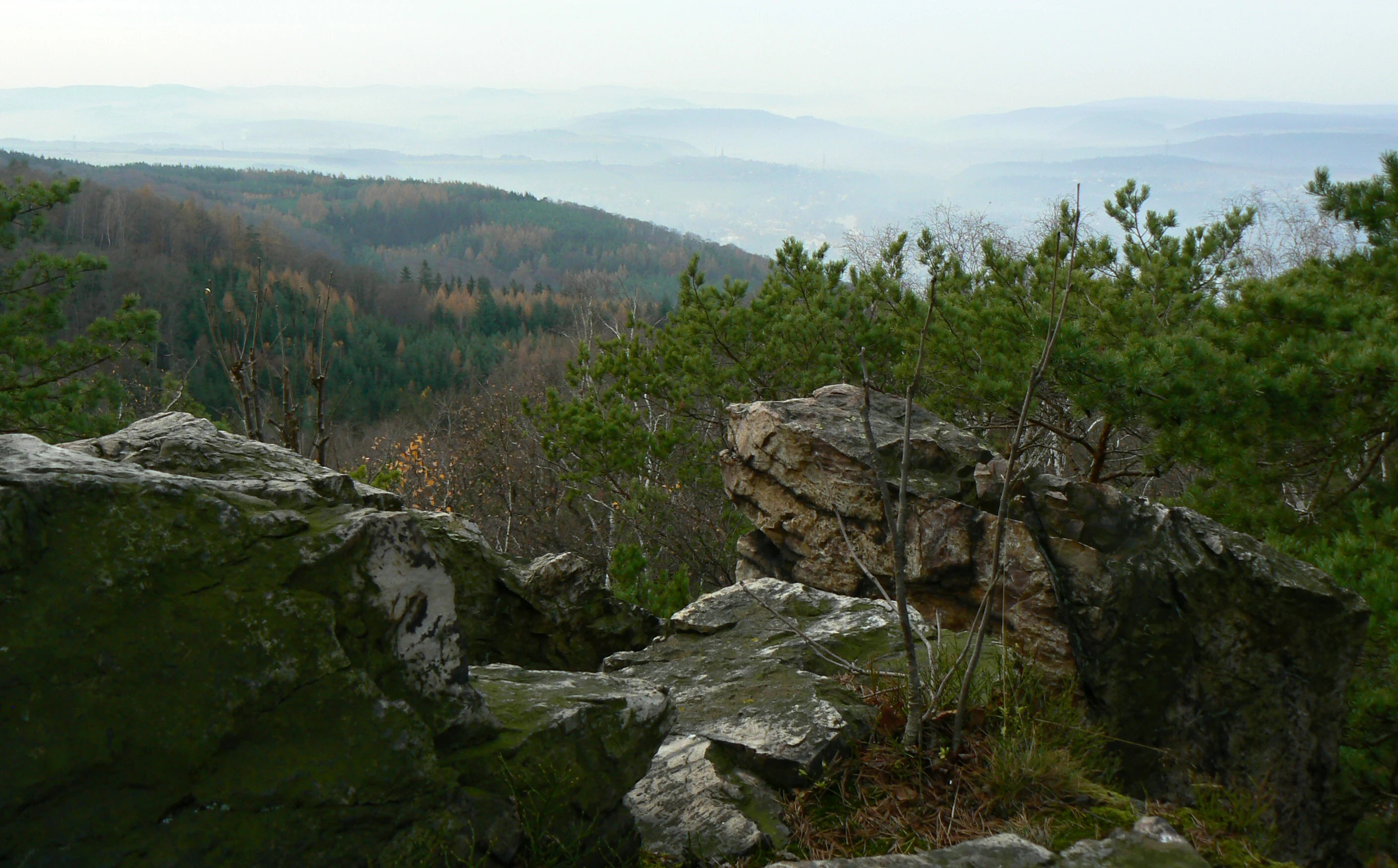 A rocky viewpoint ledge overlooking a vast, misty landscape of forested hills in the Hřebeny Nature Park.