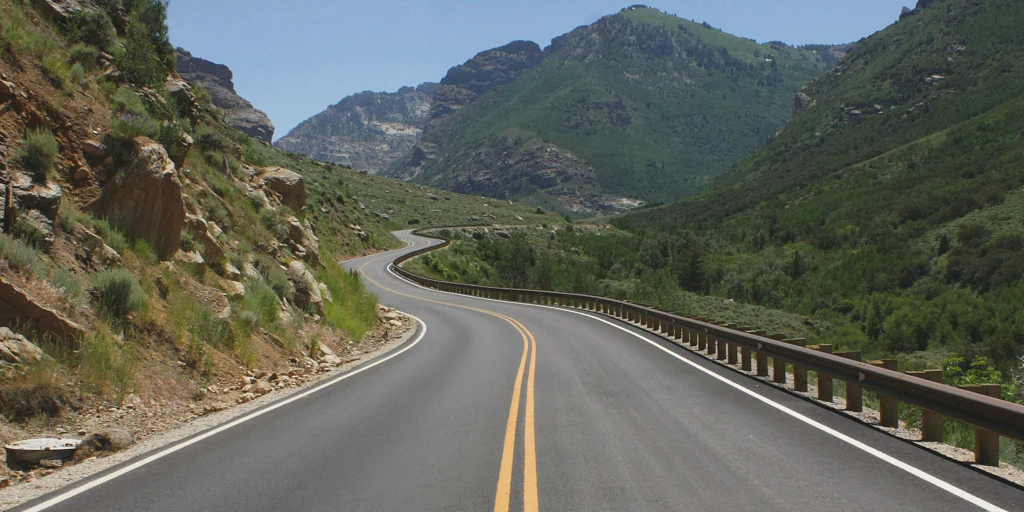 A winding asphalt road with double yellow lines travels through a steep mountain canyon; rocky hillsides are on the left and a lush green valley with mountain peaks is in the background.