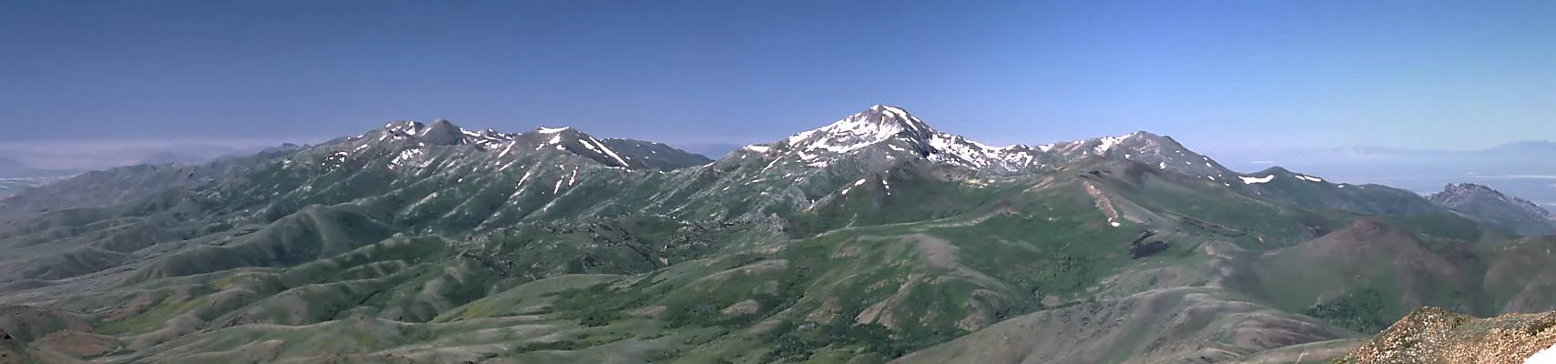 A wide panoramic view of a mountain range with patches of snow on the peaks, grassy slopes, and a clear blue sky.