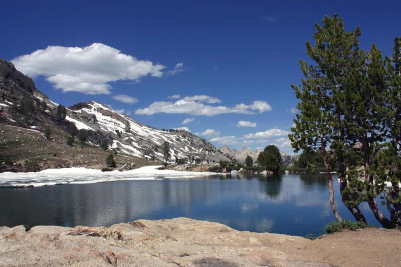 A clear blue alpine lake in the foreground, with snow-capped rocky mountains and evergreen trees in the background under a sunny blue sky.